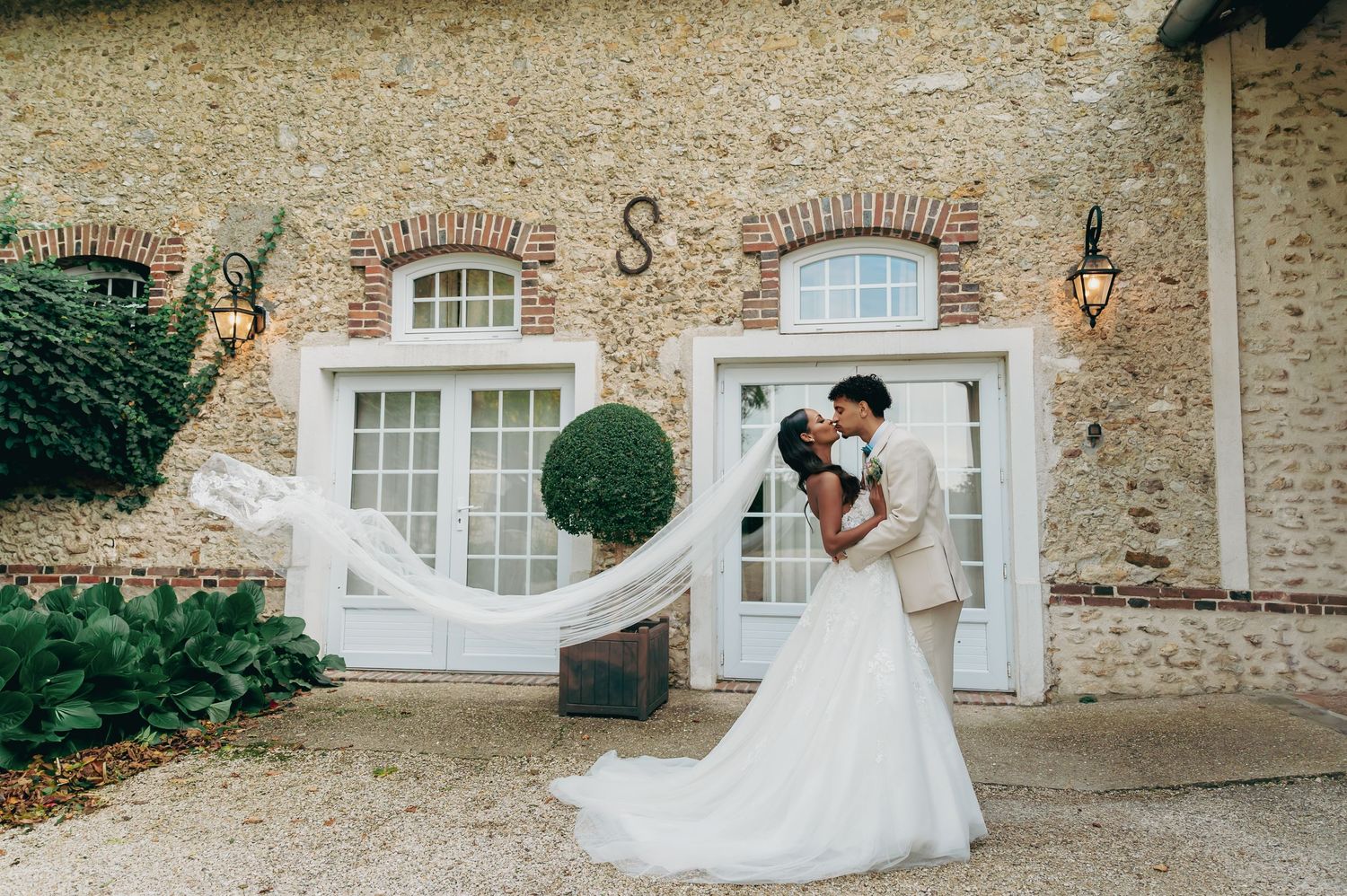 Bride and groom sharing a romantic kiss outside a rustic stone wedding venue with a flowing veil caught in the breeze.