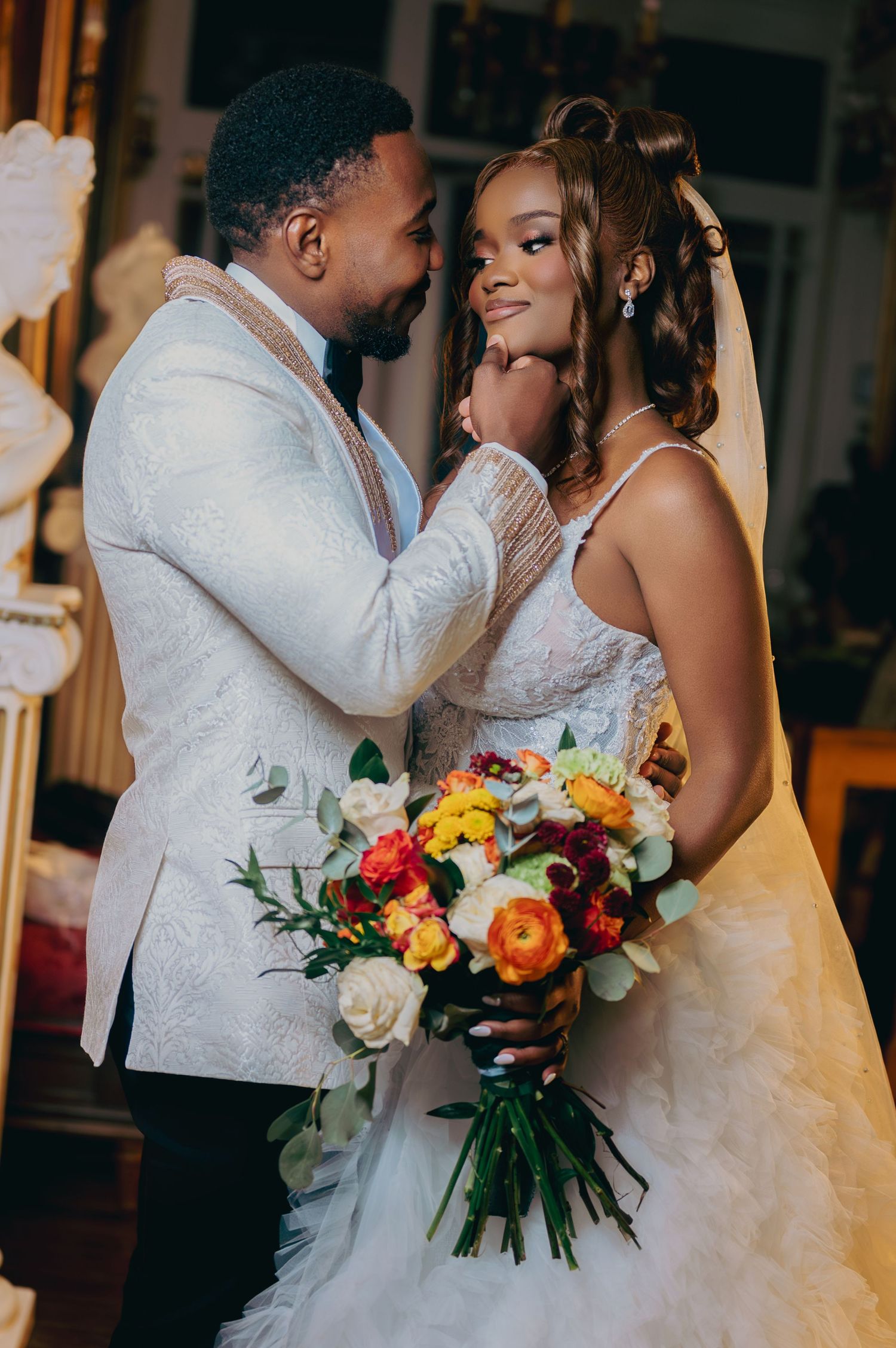 Groom tenderly holding bride's face as she smiles holding a vibrant orange and yellow bouquet.