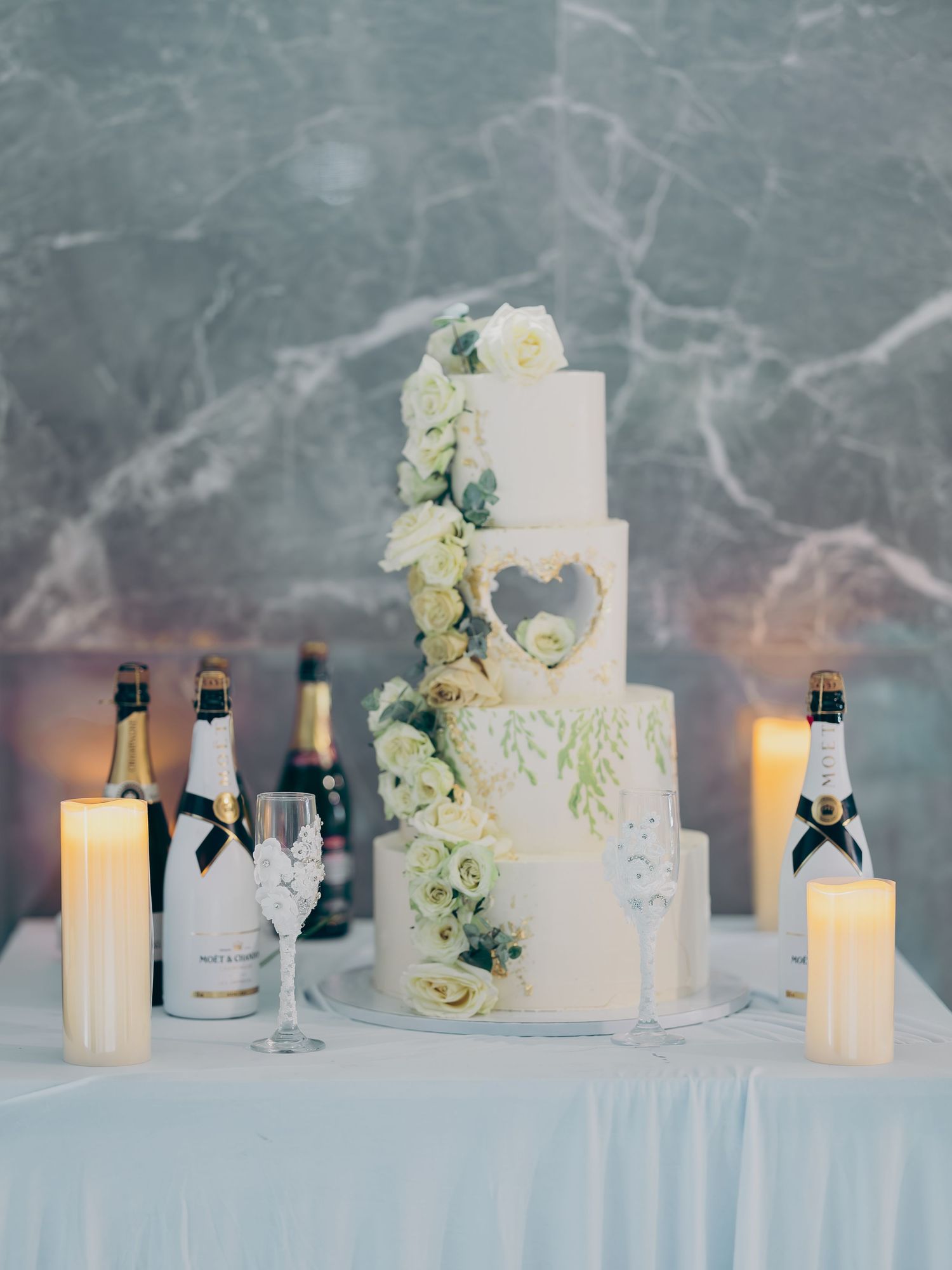 Elegant four-tier white wedding cake adorned with white roses and heart cutout flanked by champagne bottles.