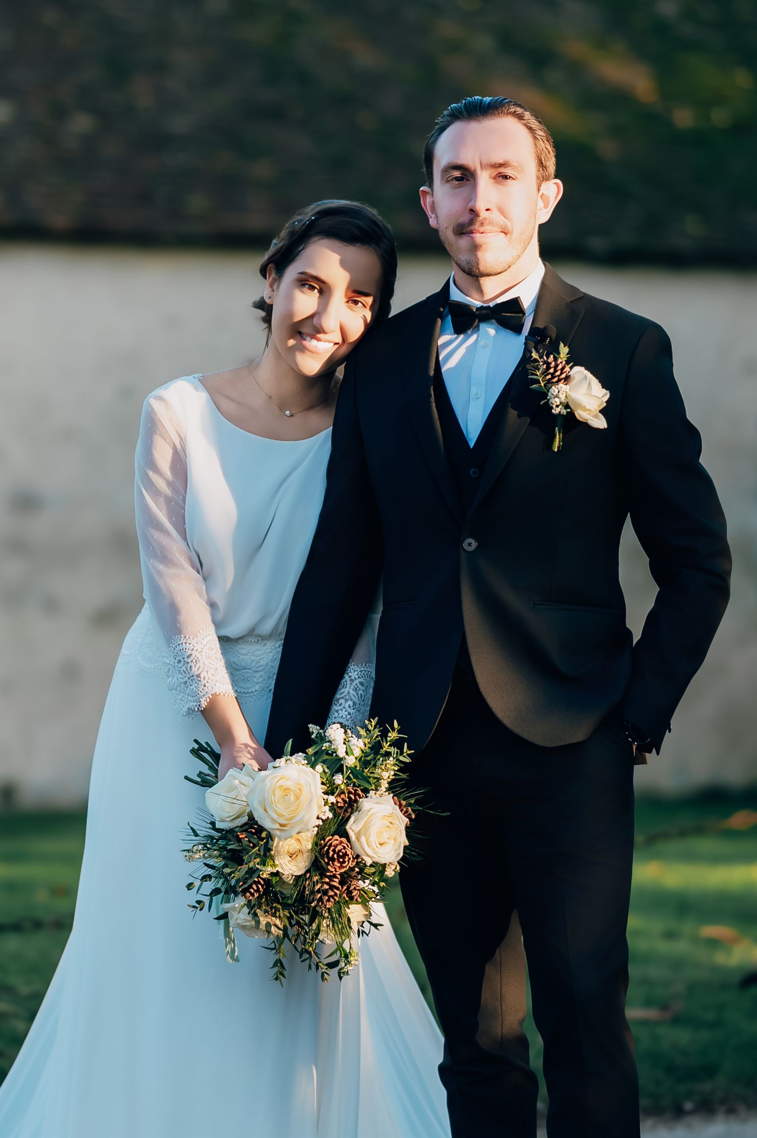 Bride in white gown holding floral bouquet stands with groom in black tuxedo at outdoor wedding.
