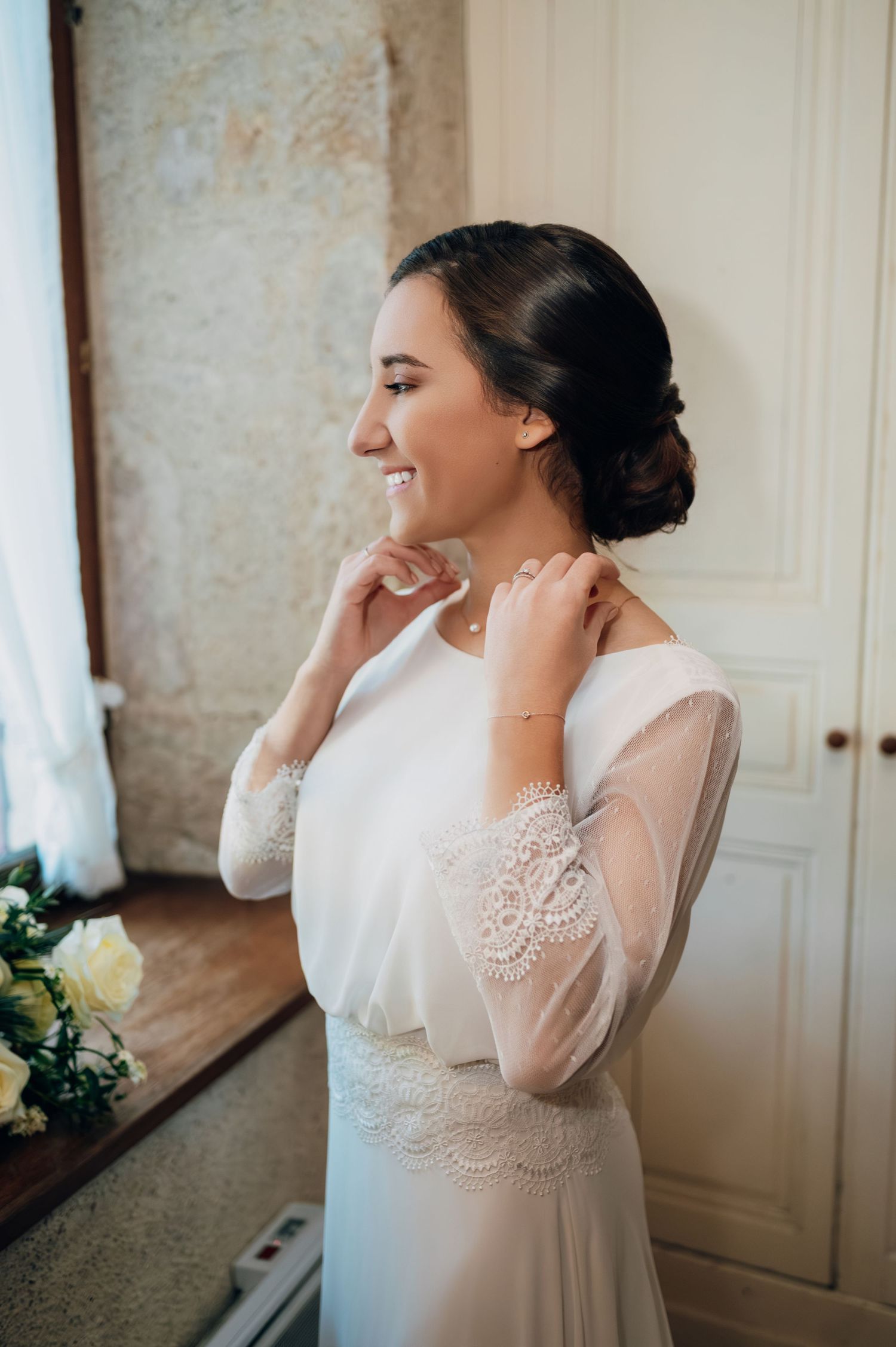 Radiant bride in long-sleeve lace wedding dress smiling while adjusting her necklace indoors.