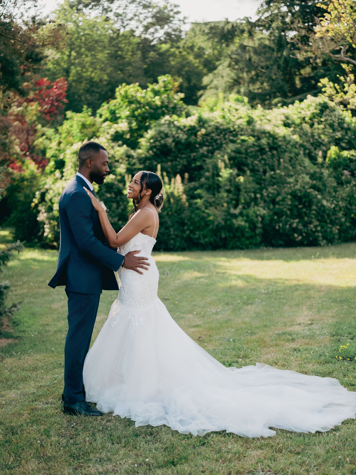 Bride in a mermaid gown with long train embraces groom in navy suit in a lush garden setting.