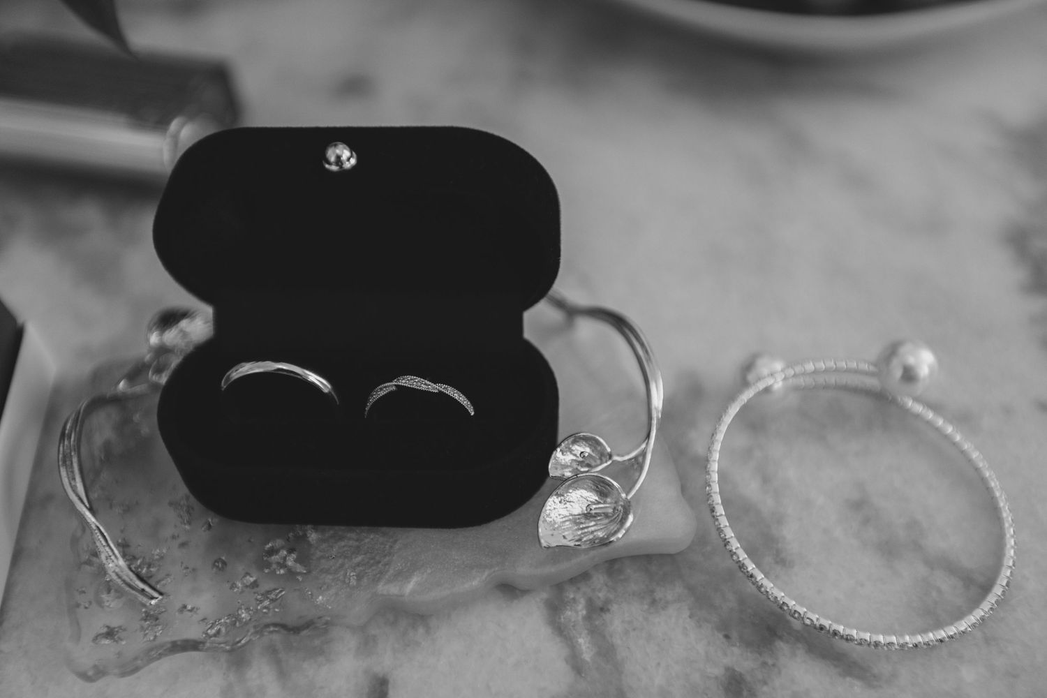 Black and white photo of two wedding rings in velvet box beside silver bracelet.