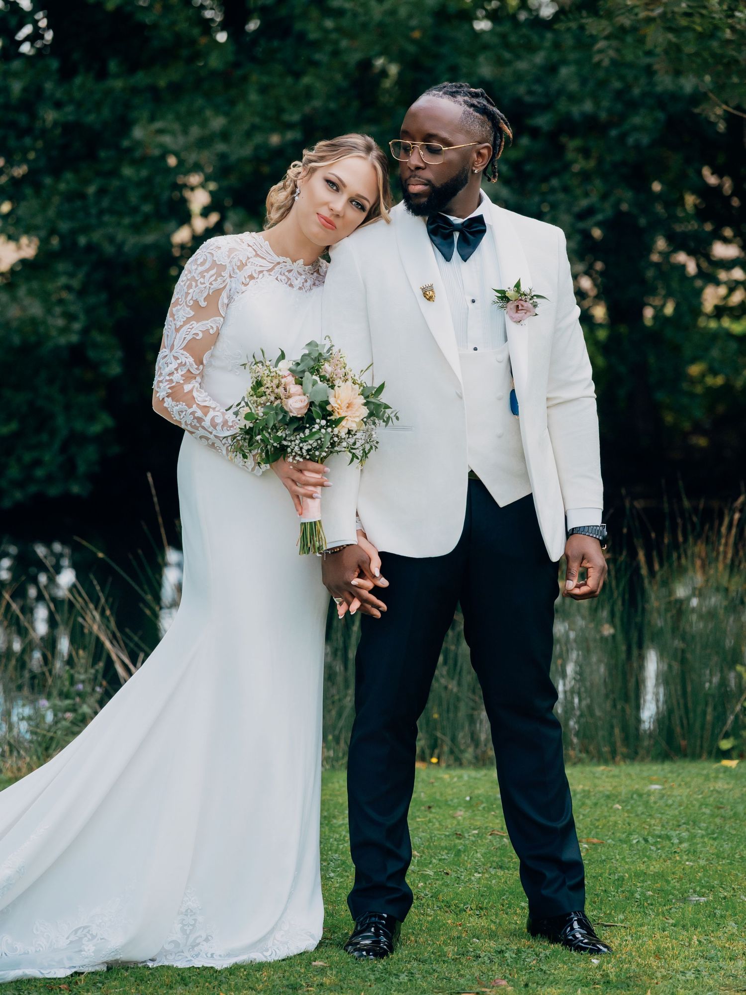 Bride in long-sleeve lace wedding dress and groom in white tuxedo posing together in a garden.