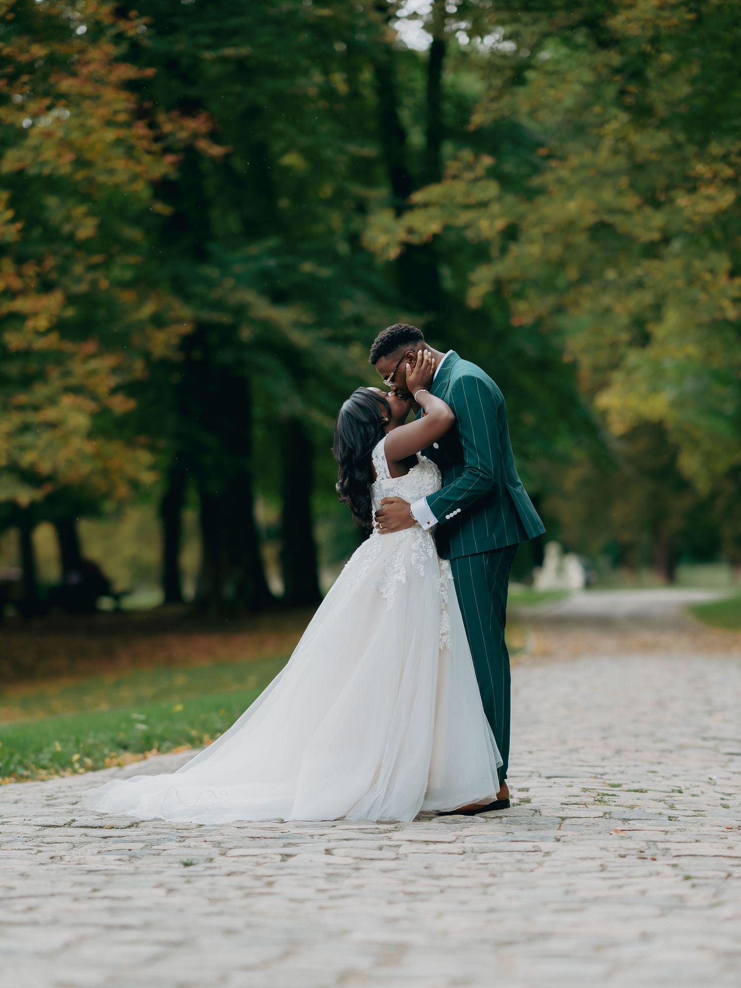Newlywed couple sharing a romantic kiss on a cobblestone path surrounded by autumn trees.