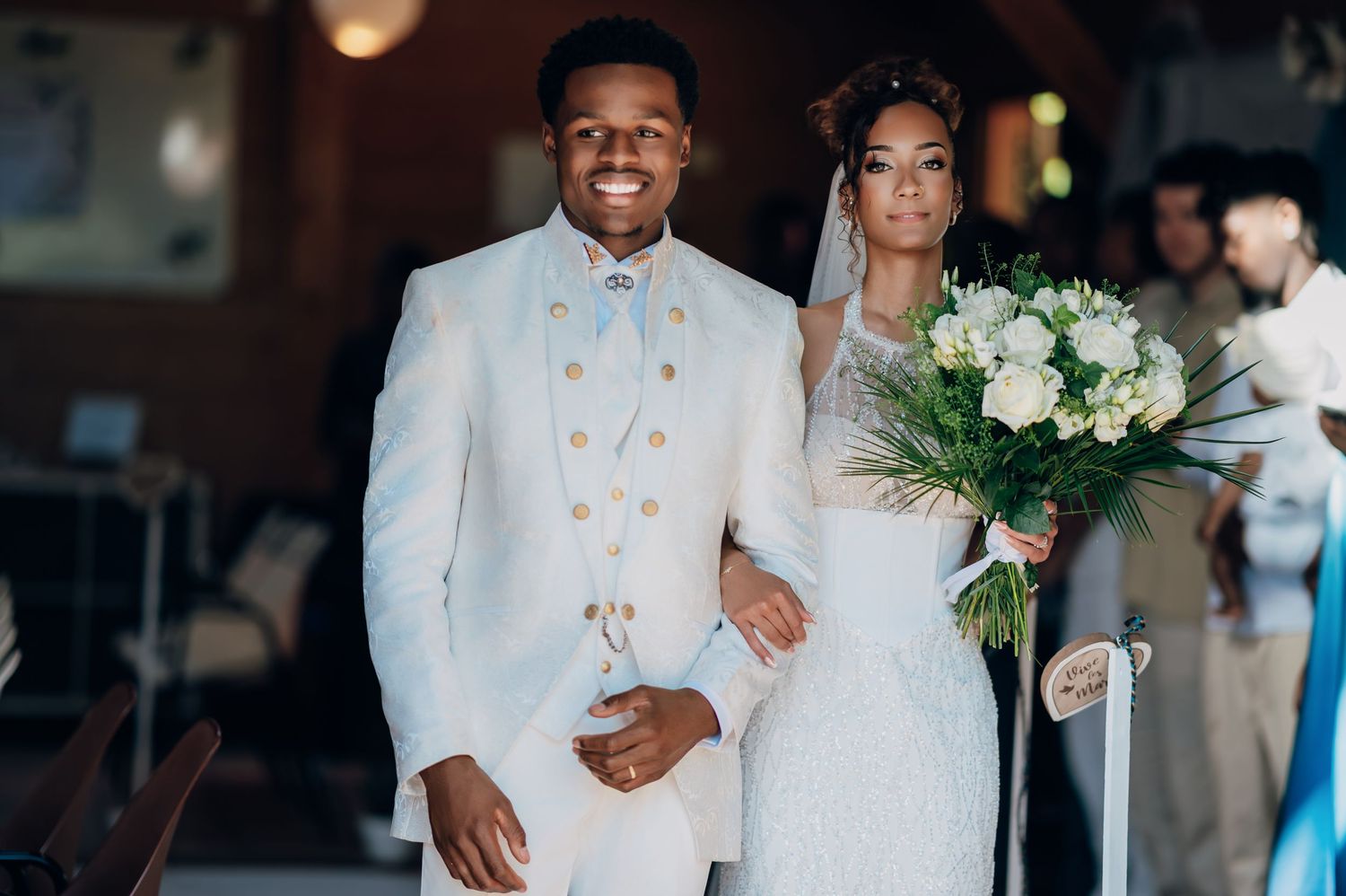Smiling groom in white military-style suit escorts bride in lace gown down the aisle holding white bouquet.