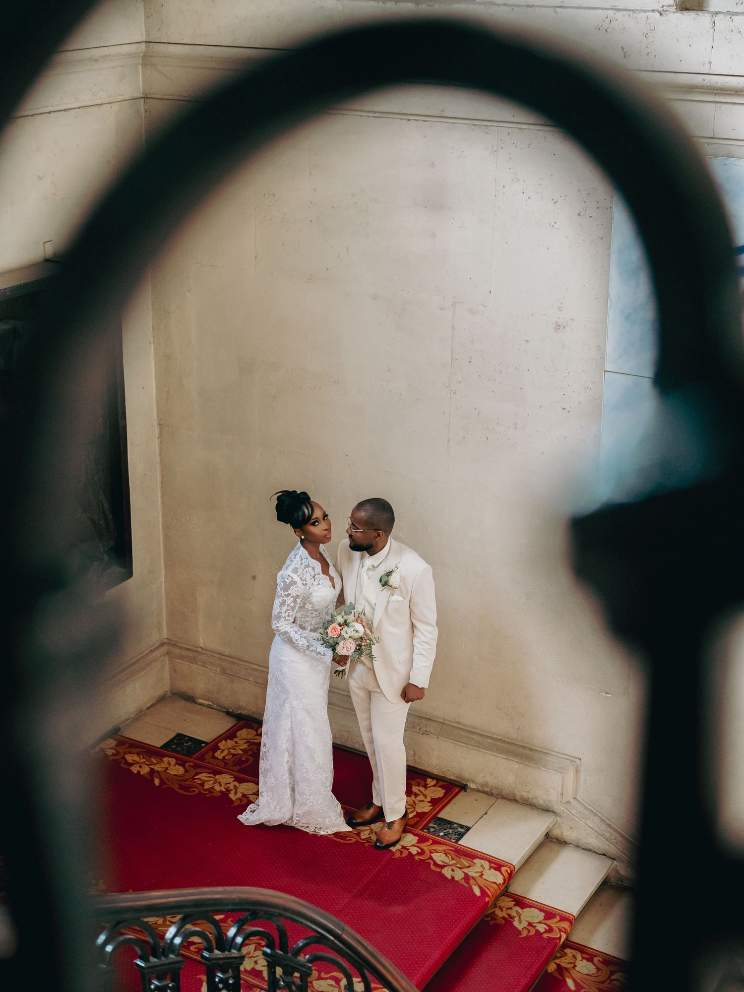 Wedding couple shares romantic moment on red carpet staircase viewed through ornate iron railing.