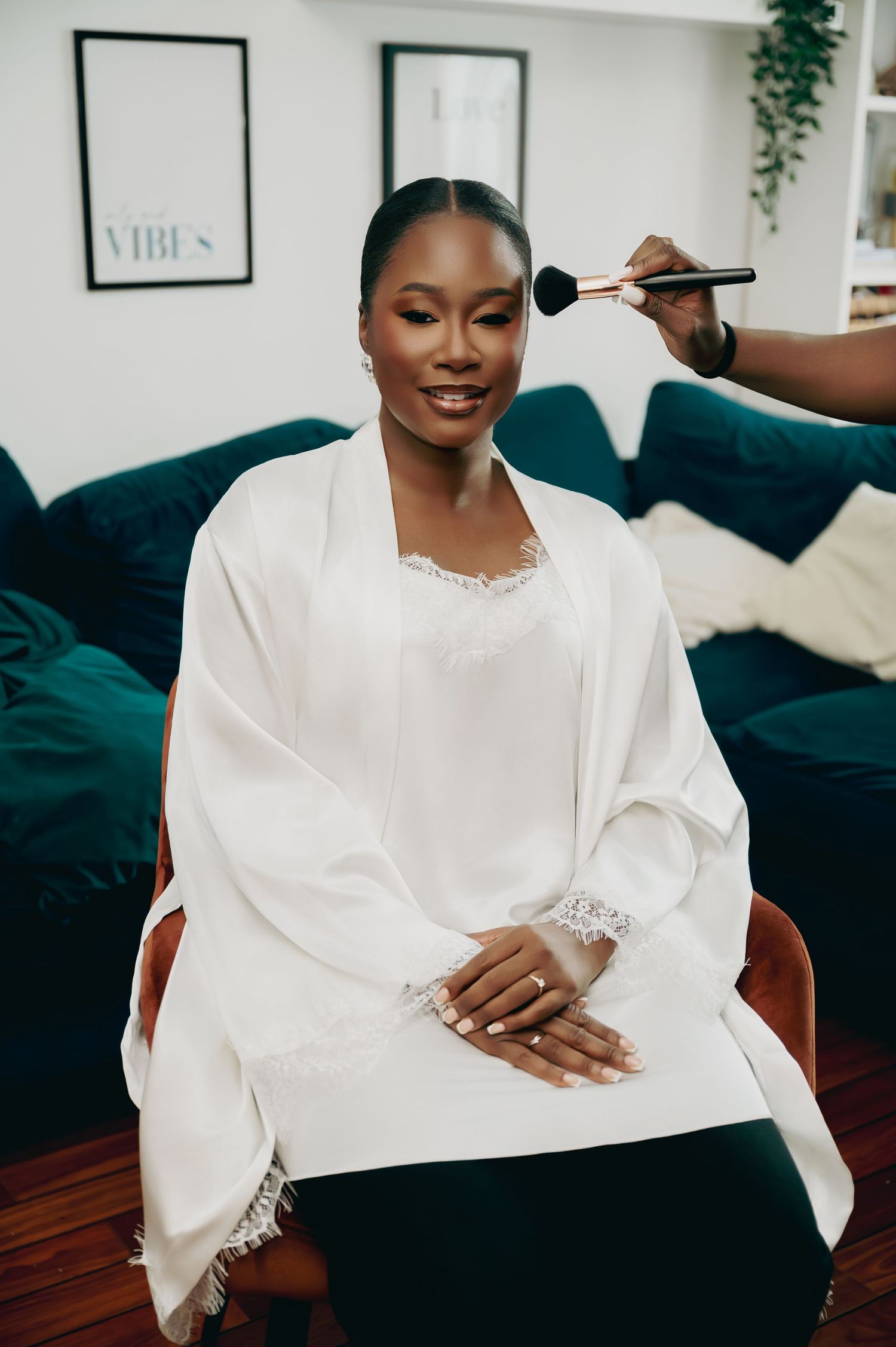 Bride in white lace robe smiles warmly as a makeup artist applies blush during bridal preparations.