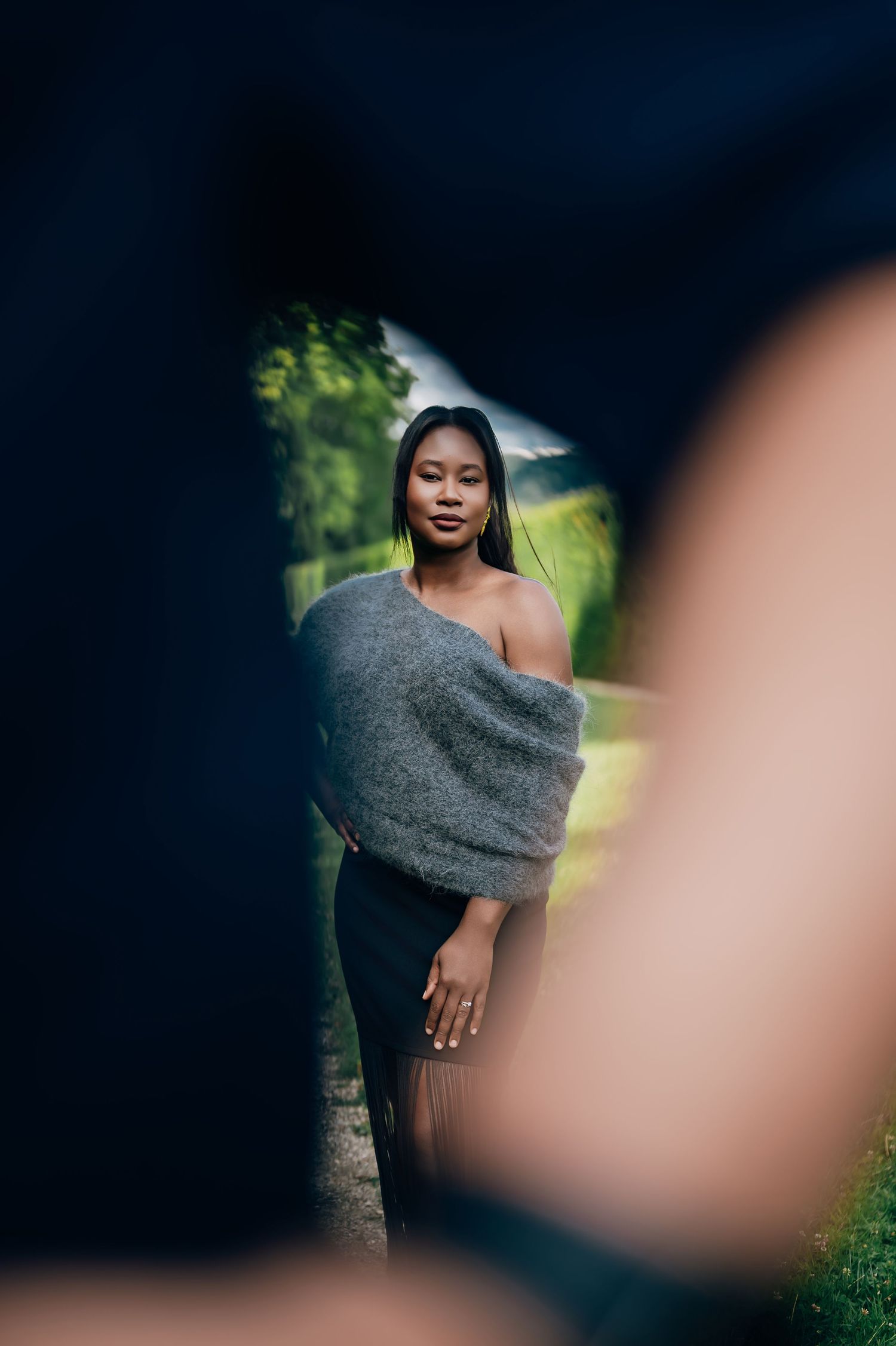 Young woman in gray off-shoulder wrap and black skirt stands on garden path, framed by dark foreground elements.