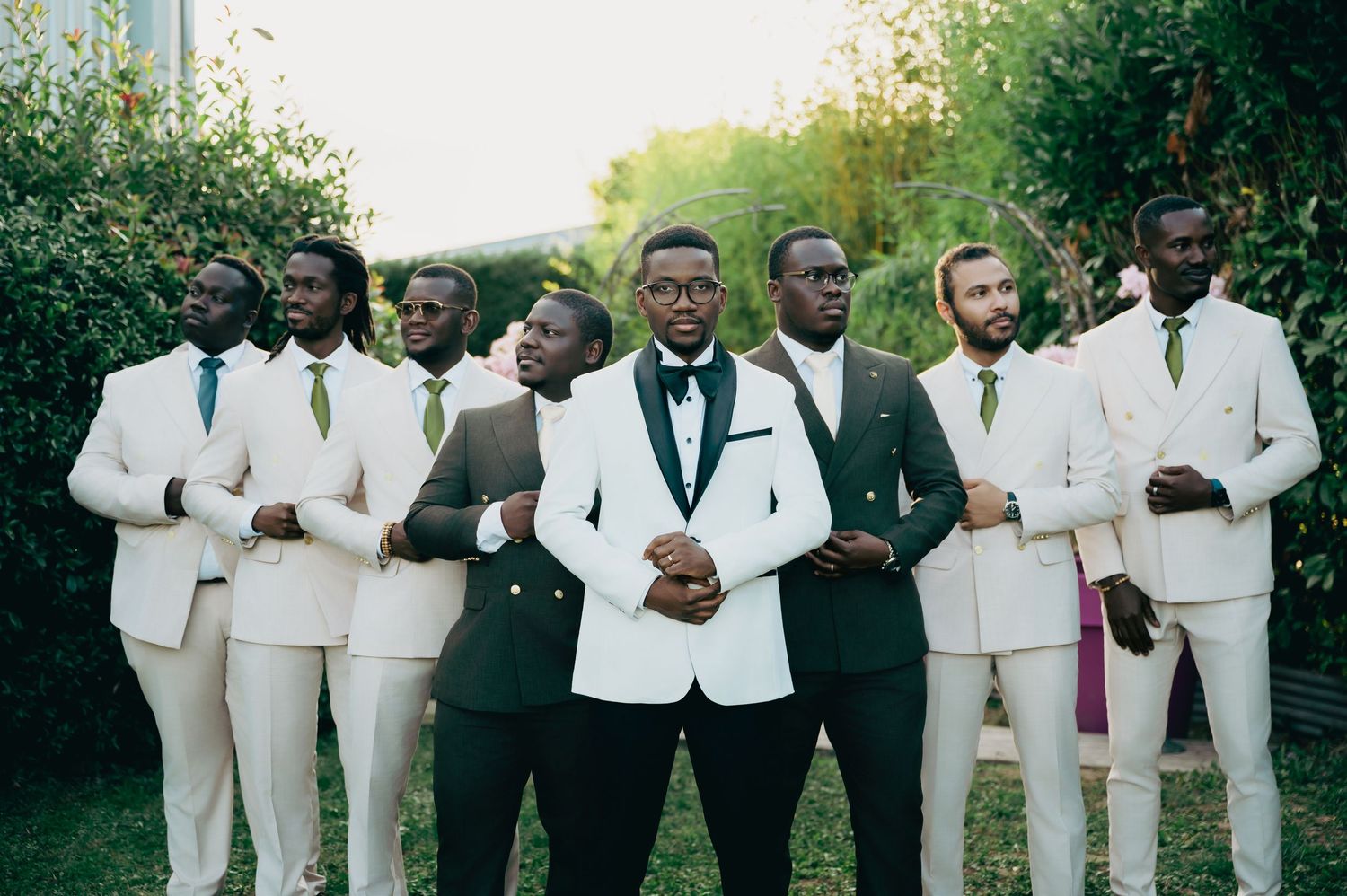 Groom in white tuxedo stands with groomsmen in green and white suits for formal portrait.
