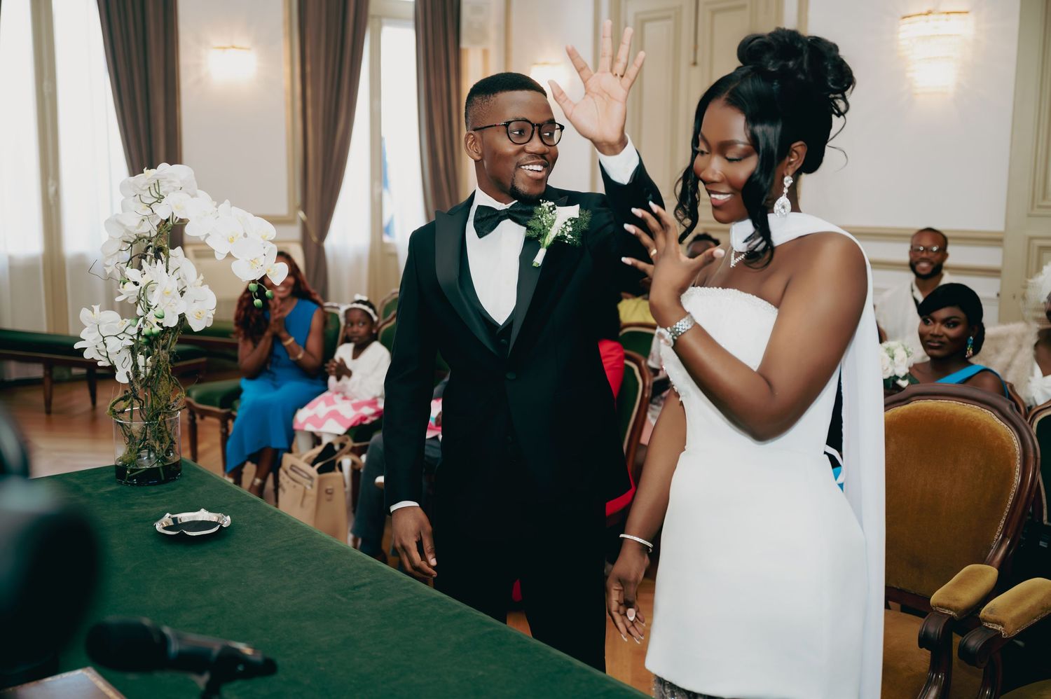 Joyful bride and groom wave to guests during wedding ceremony at elegant green-draped altar.