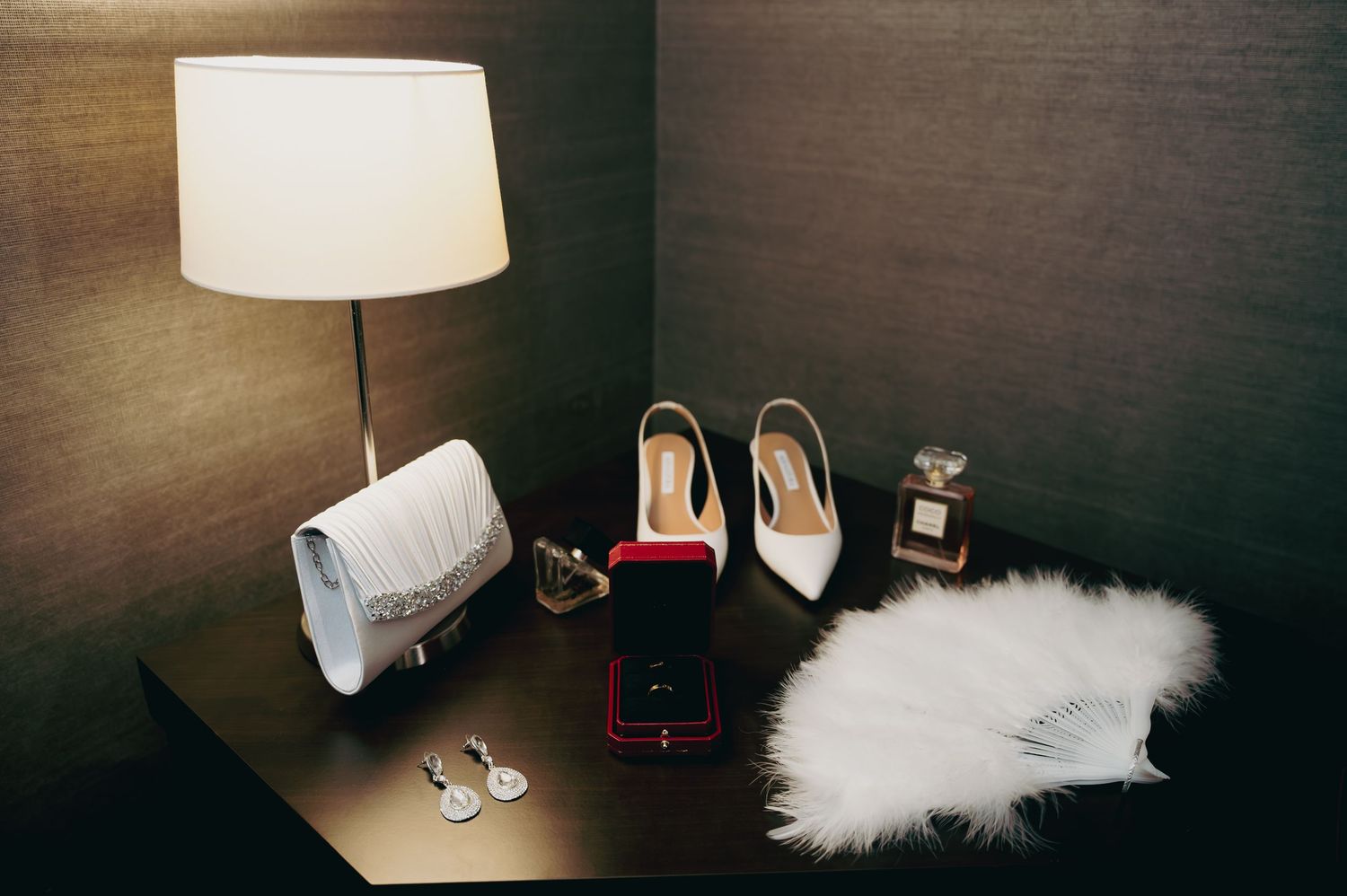 Bridal flat lay with white heels, clutch, perfume, earrings, and ring box on a dark table.