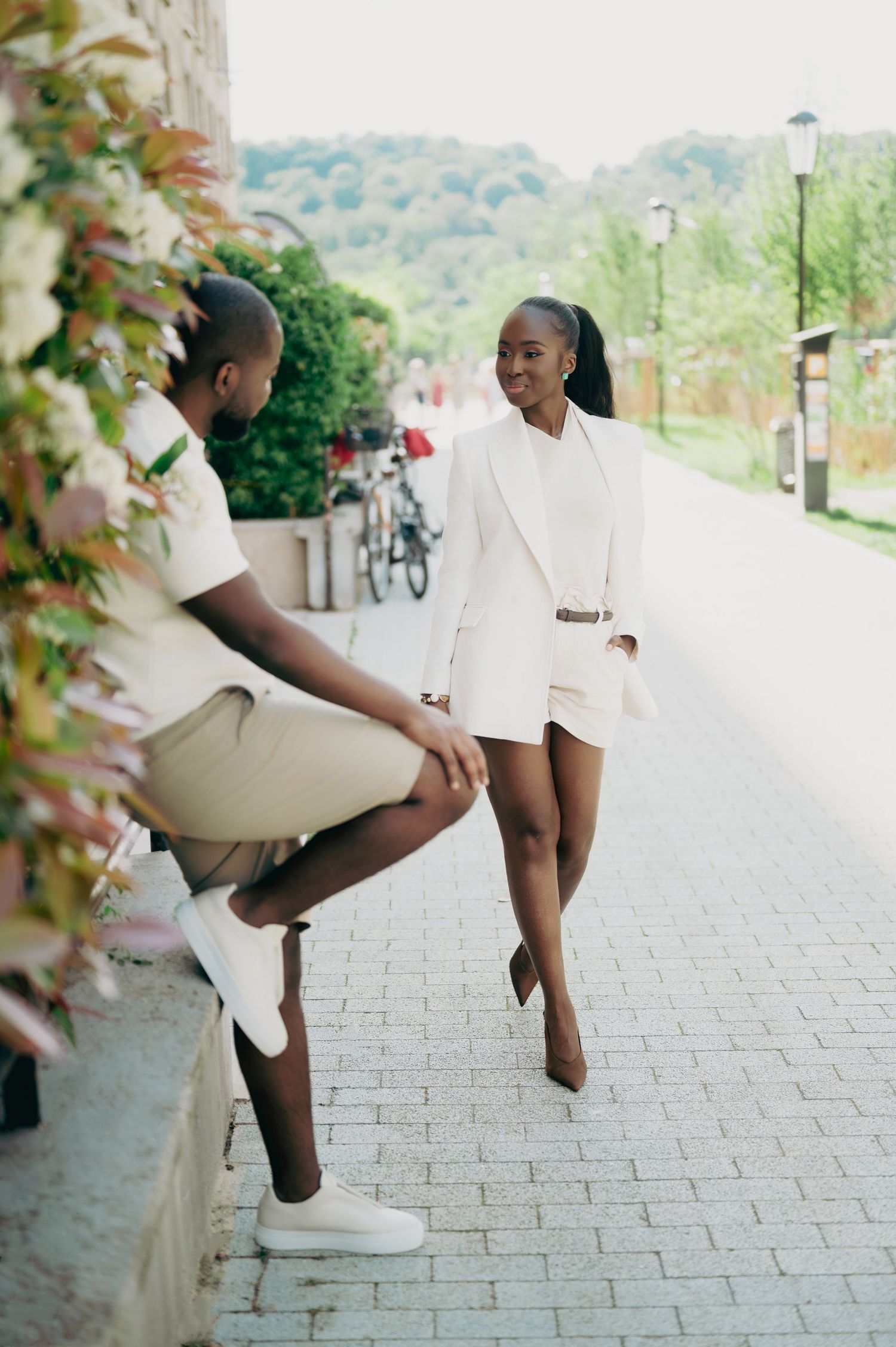 Smiling Black couple in coordinated white and beige outfits interact on a charming outdoor walkway.