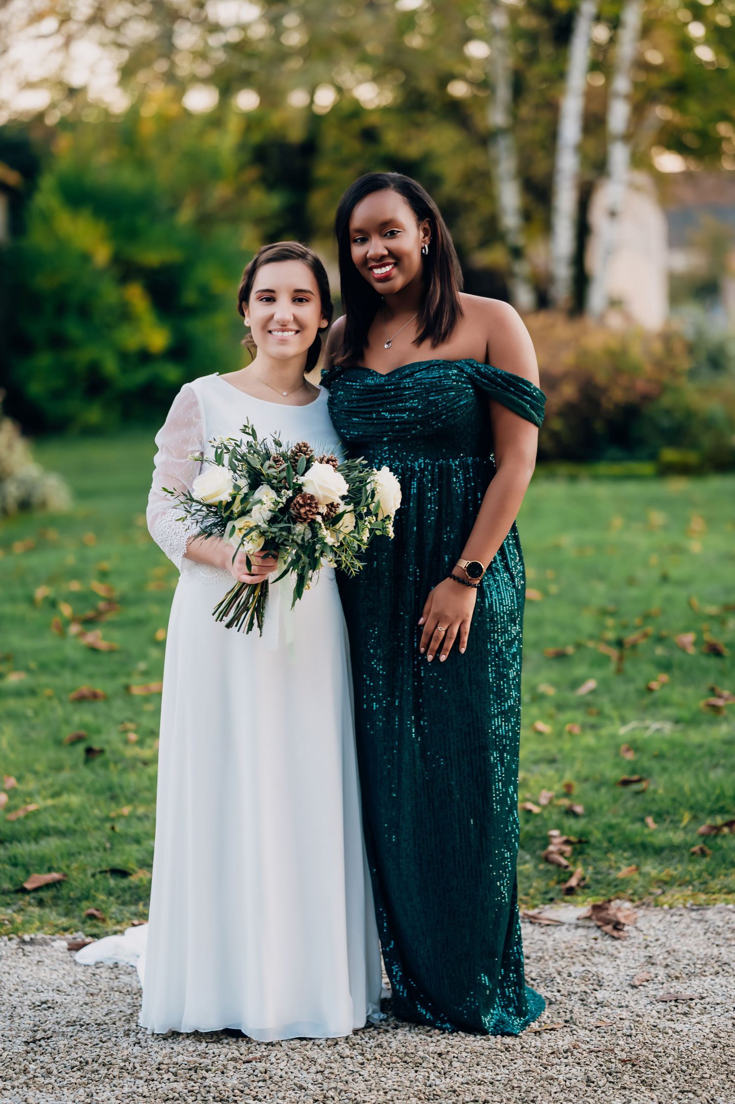 Bride in white lace gown poses with bridesmaid wearing emerald green sequin off-shoulder dress outdoors.
