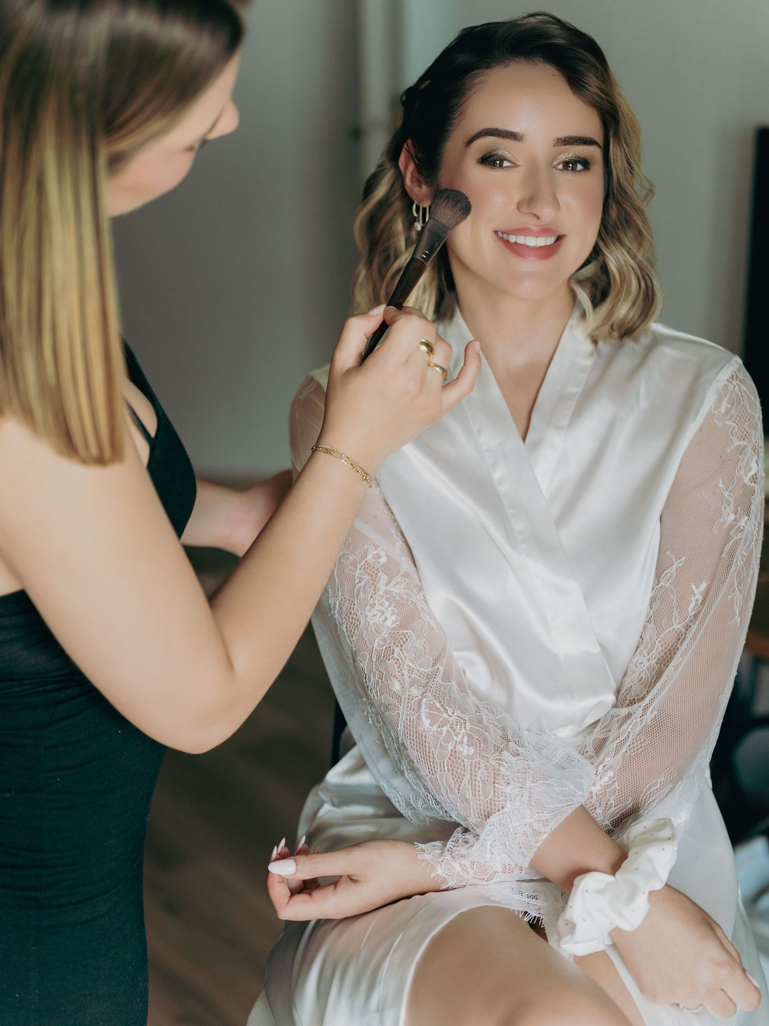 A bride smiles as a makeup artist applies finishing touches during wedding day preparations.