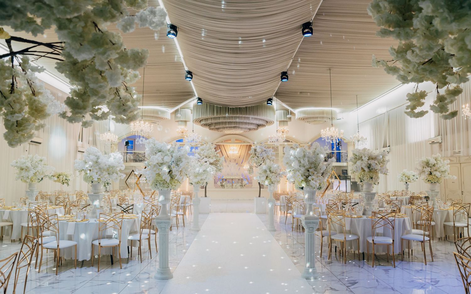 Breathtaking white wedding reception hall with floral ceiling installations, gold chairs, and a glowing chandelier aisle.