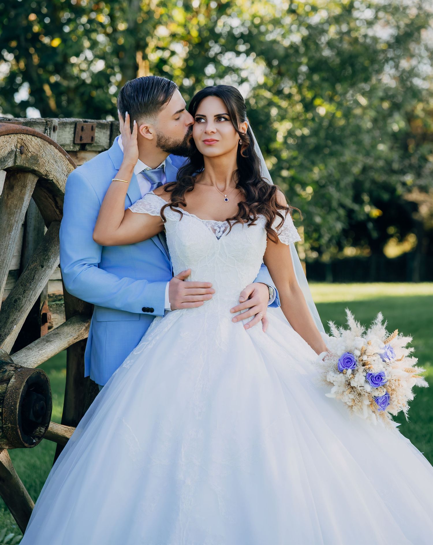 Groom in light blue suit kissing bride's cheek as she holds bouquet near rustic wooden wagon wheel outdoors.