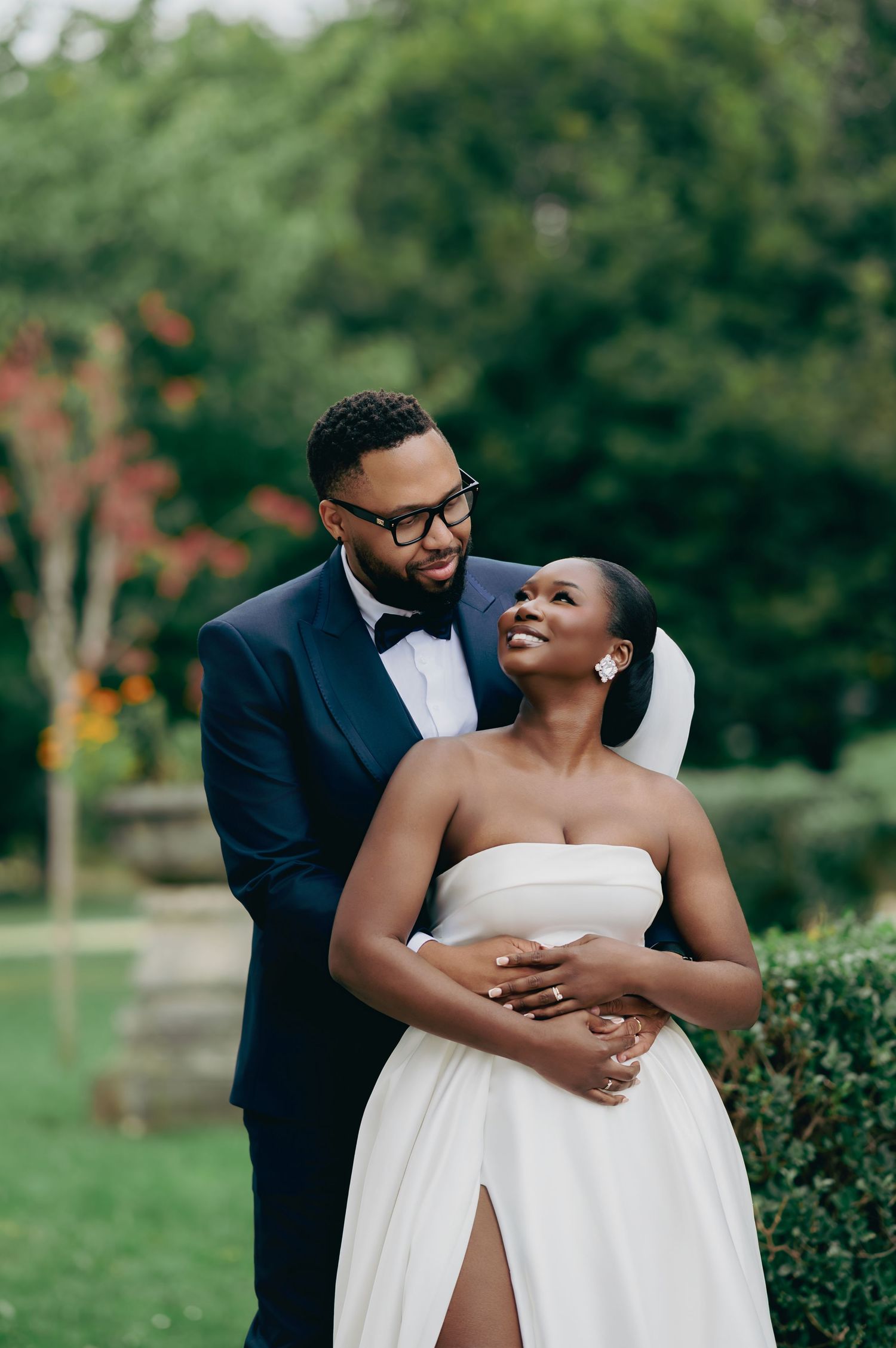 Groom in navy tuxedo embraces smiling bride in strapless white gown with slit in a garden setting.