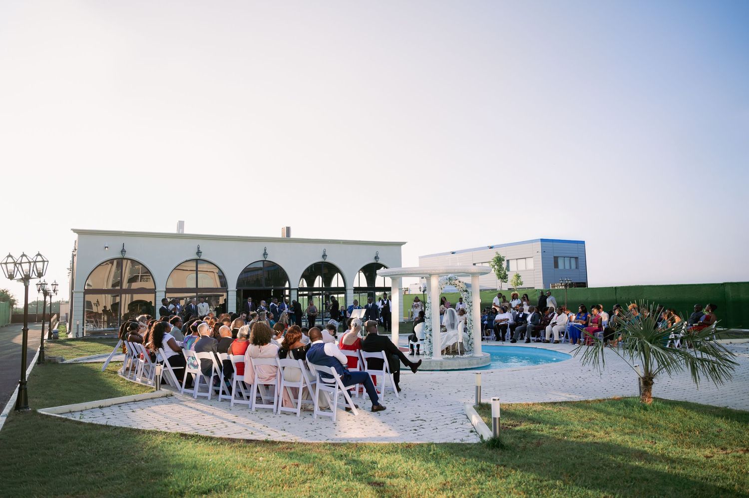 Outdoor wedding ceremony by a pool at a white villa venue at dusk, with guests seated in white chairs.