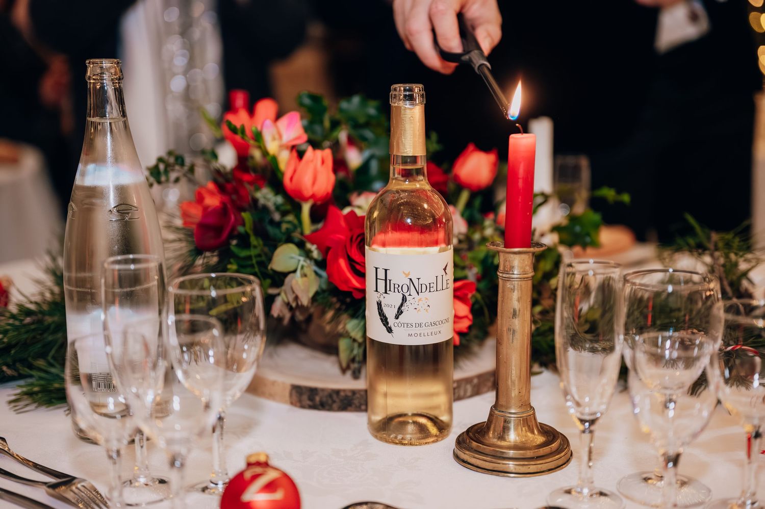 Hand lighting a red candle beside a wine bottle and red floral centerpiece at a Christmas wedding reception table.