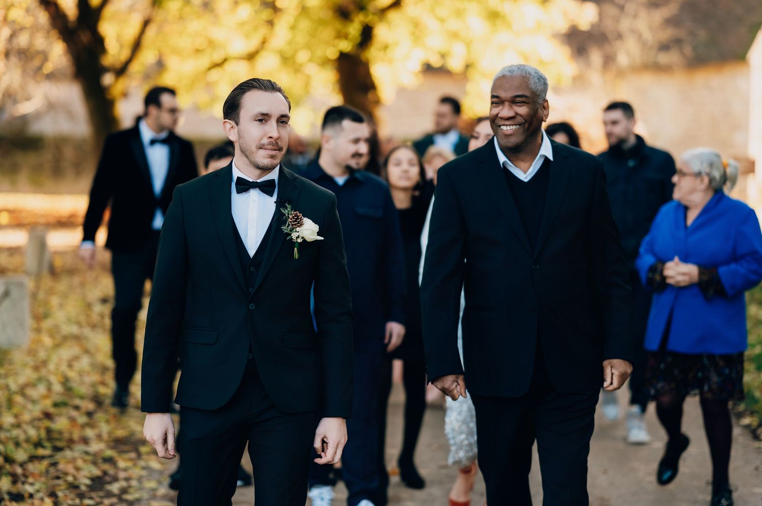 Two men in black suits walking together along an autumn tree-lined path during a wedding celebration outdoors.