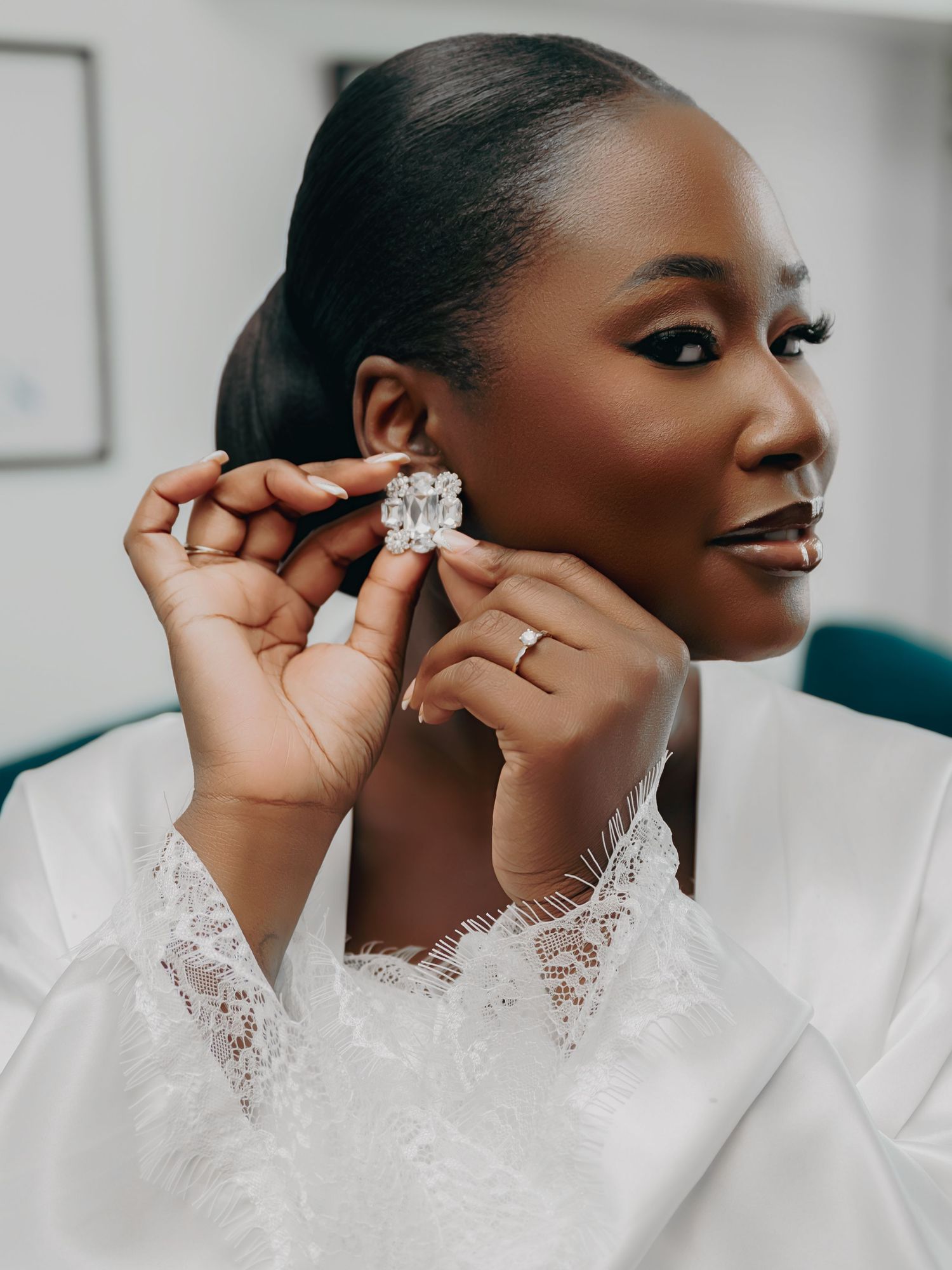 Bride in white lace robe puts on a large crystal earring while showcasing her engagement ring.