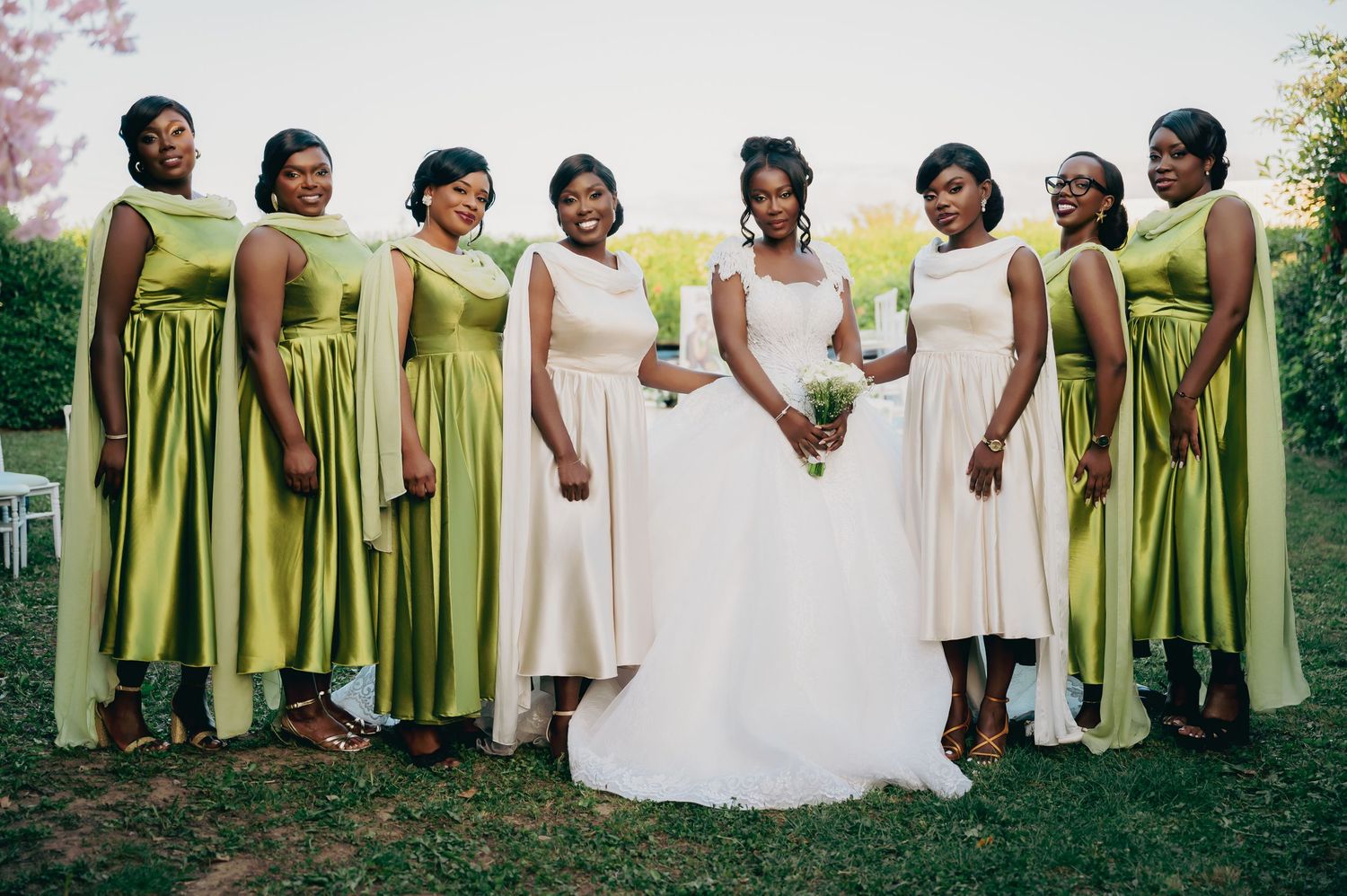 Bride in white gown poses with seven bridesmaids in olive green dresses outdoors.