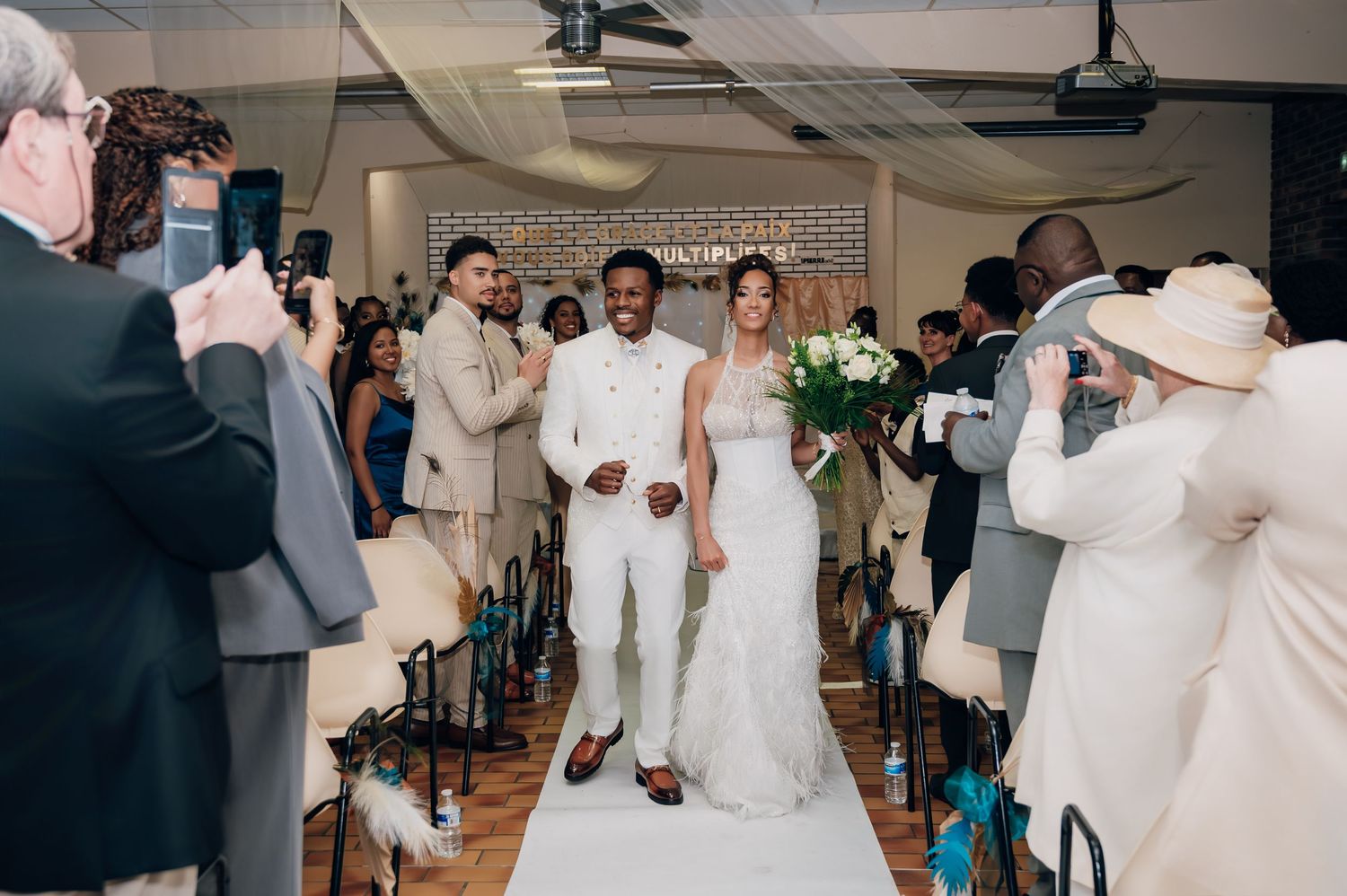 Interracial couple walks down wedding aisle in white outfits as guests celebrate and photograph the ceremony.