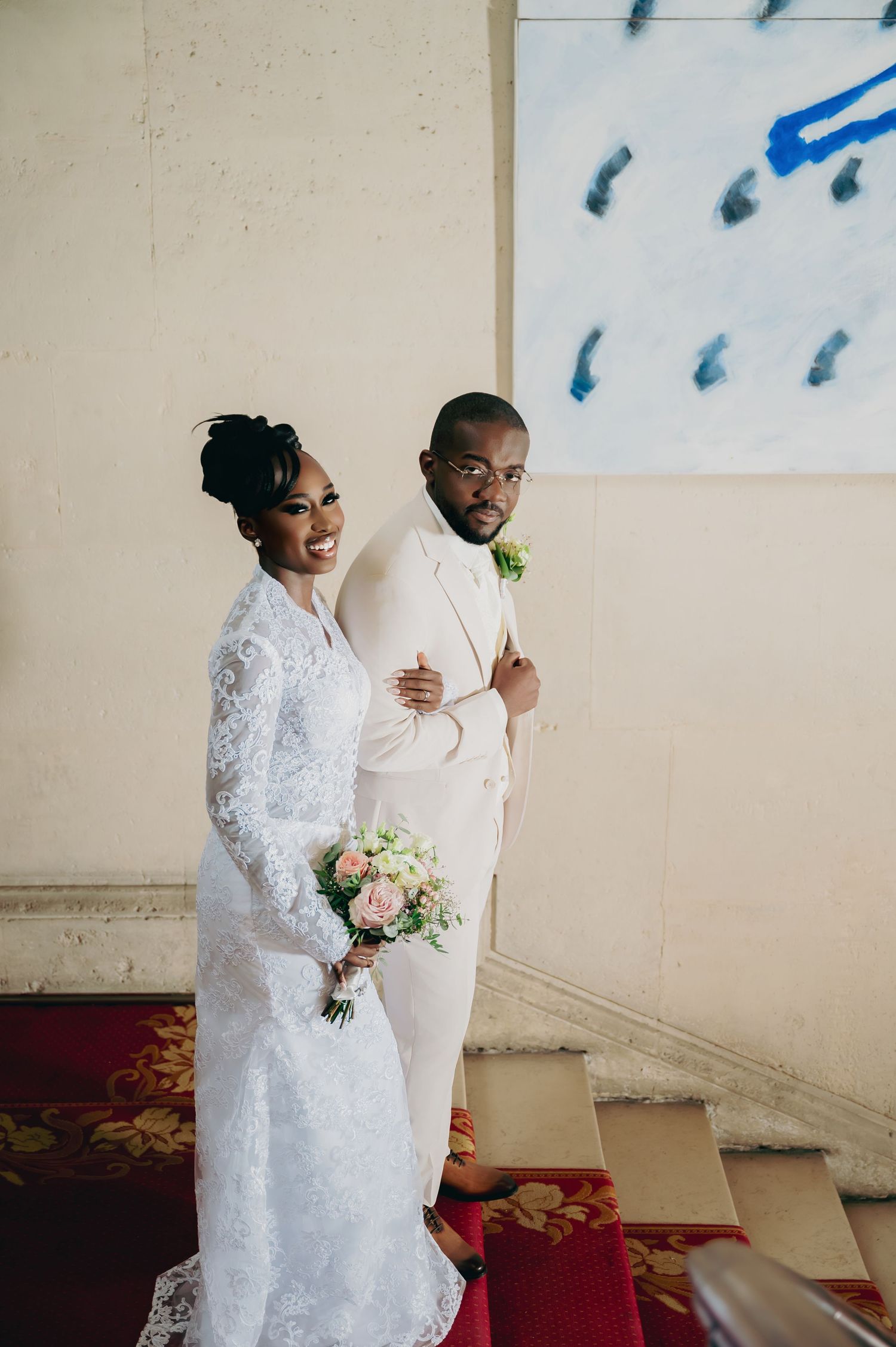 Bride in lace gown holds bouquet beside groom in cream suit at elegant wedding venue.