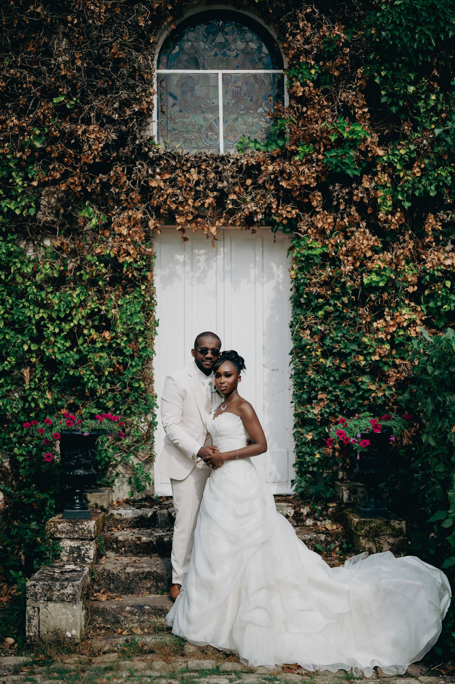 Bride and groom pose together before ivy-covered stone wall with white arched door and pink flowers.
