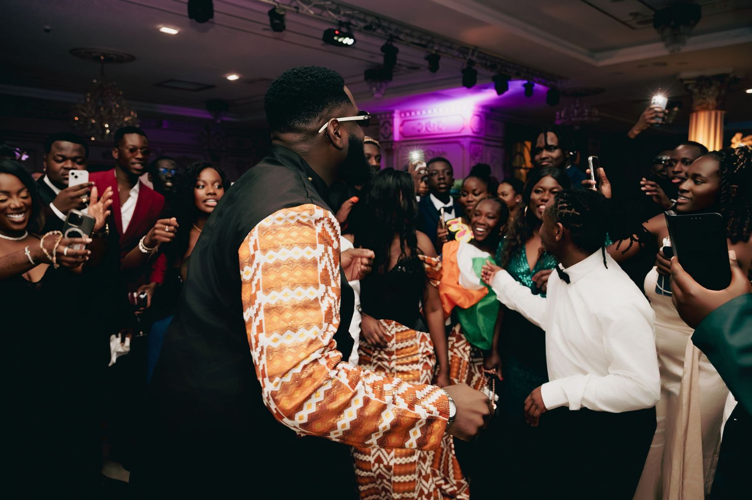 Groom in traditional African kente jacket dancing energetically with guests at a wedding reception.