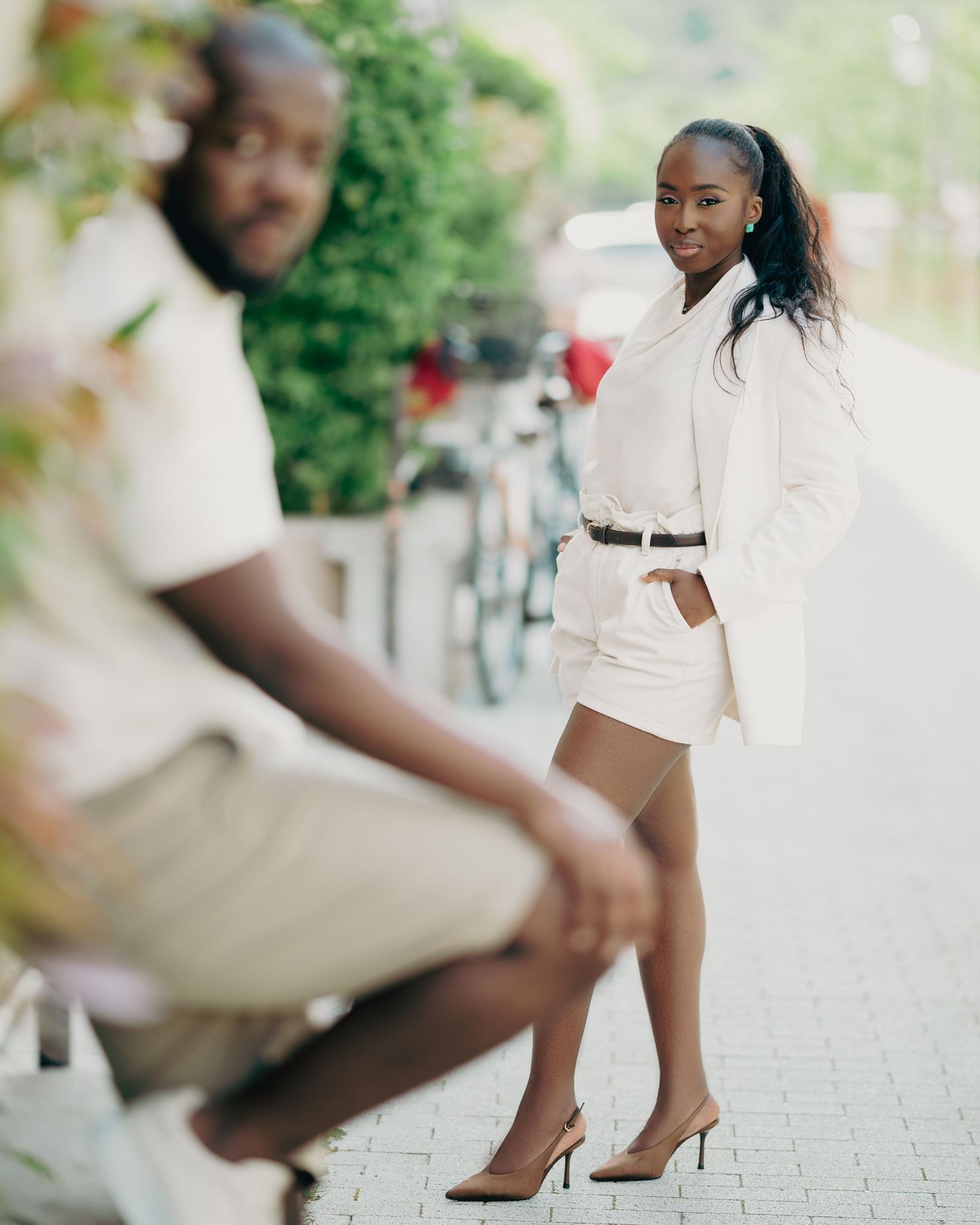 Stylish Black woman in white blazer dress and heels poses outdoors while man sits blurred in foreground.