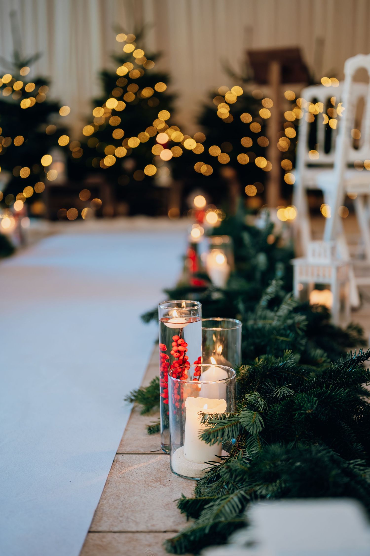 Close-up of Christmas wedding aisle decor featuring glass candle holders, red berries, and evergreen garland with bokeh tree lights.