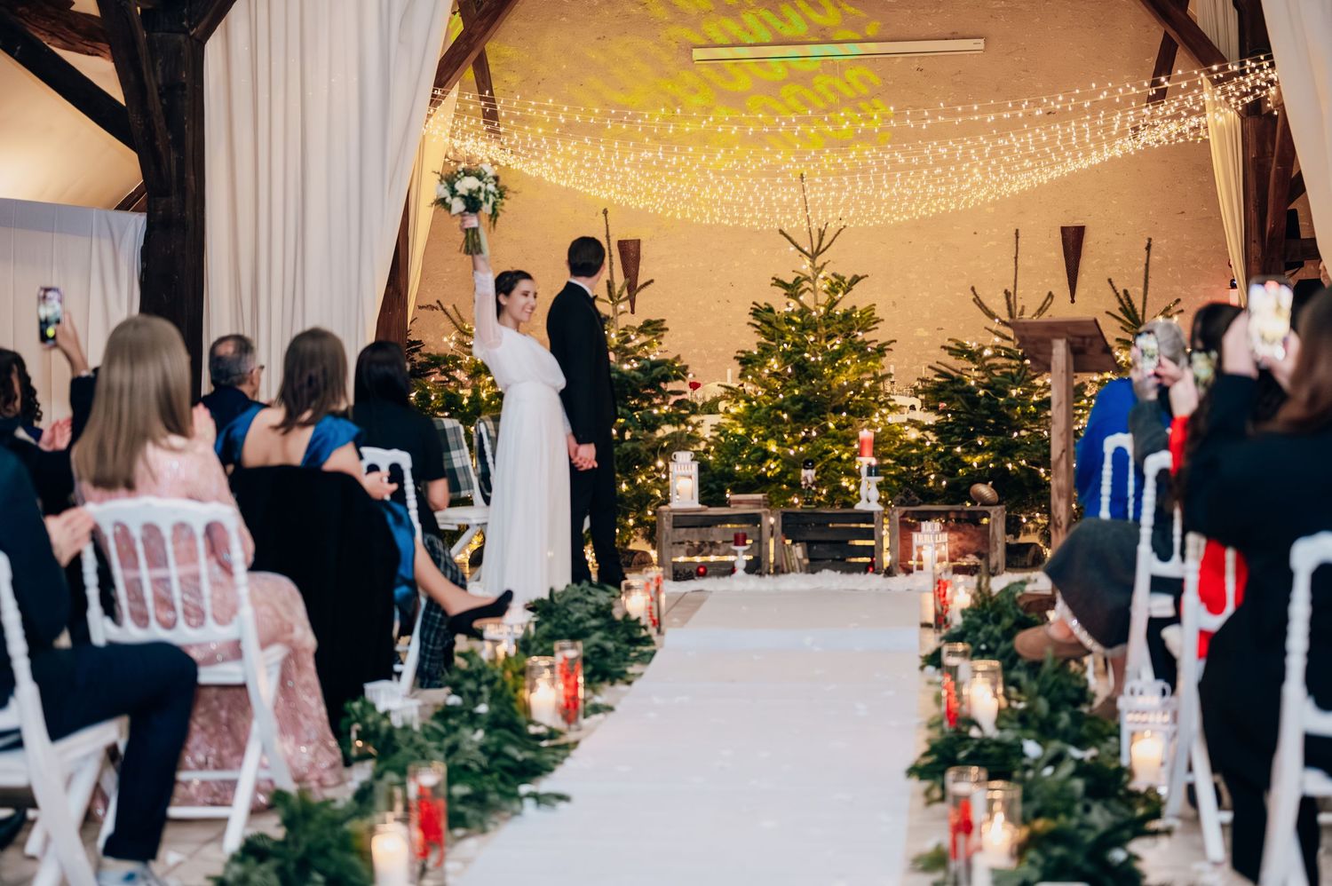 Winter wedding ceremony in a rustic barn with white aisle runner, greenery garlands, lanterns, and fairy lights overhead.