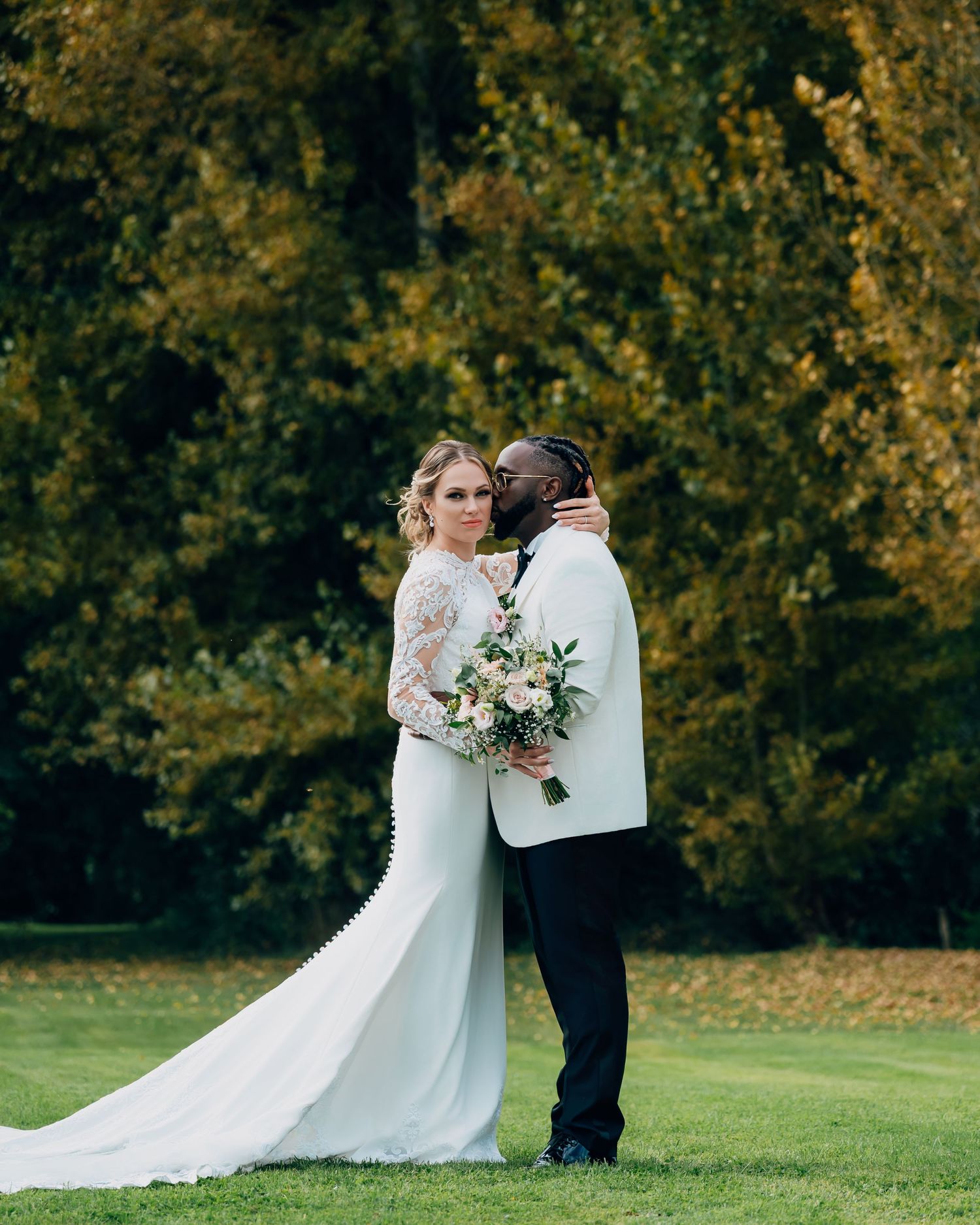Bride in long-sleeve lace gown and groom in white tuxedo embracing outdoors with autumn trees behind.