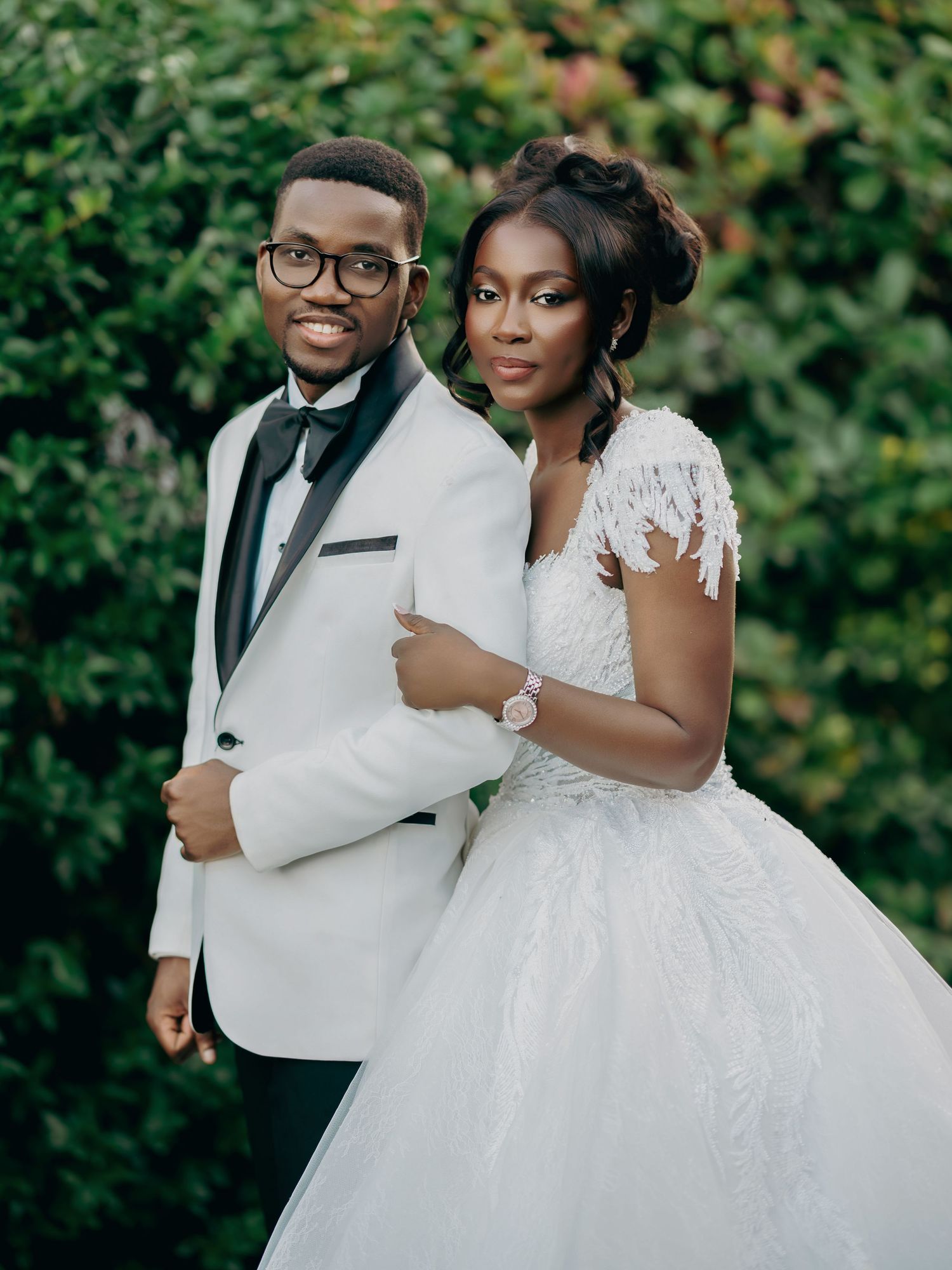 Black couple in wedding attire pose outdoors, groom in white tuxedo and bride in lace ball gown.