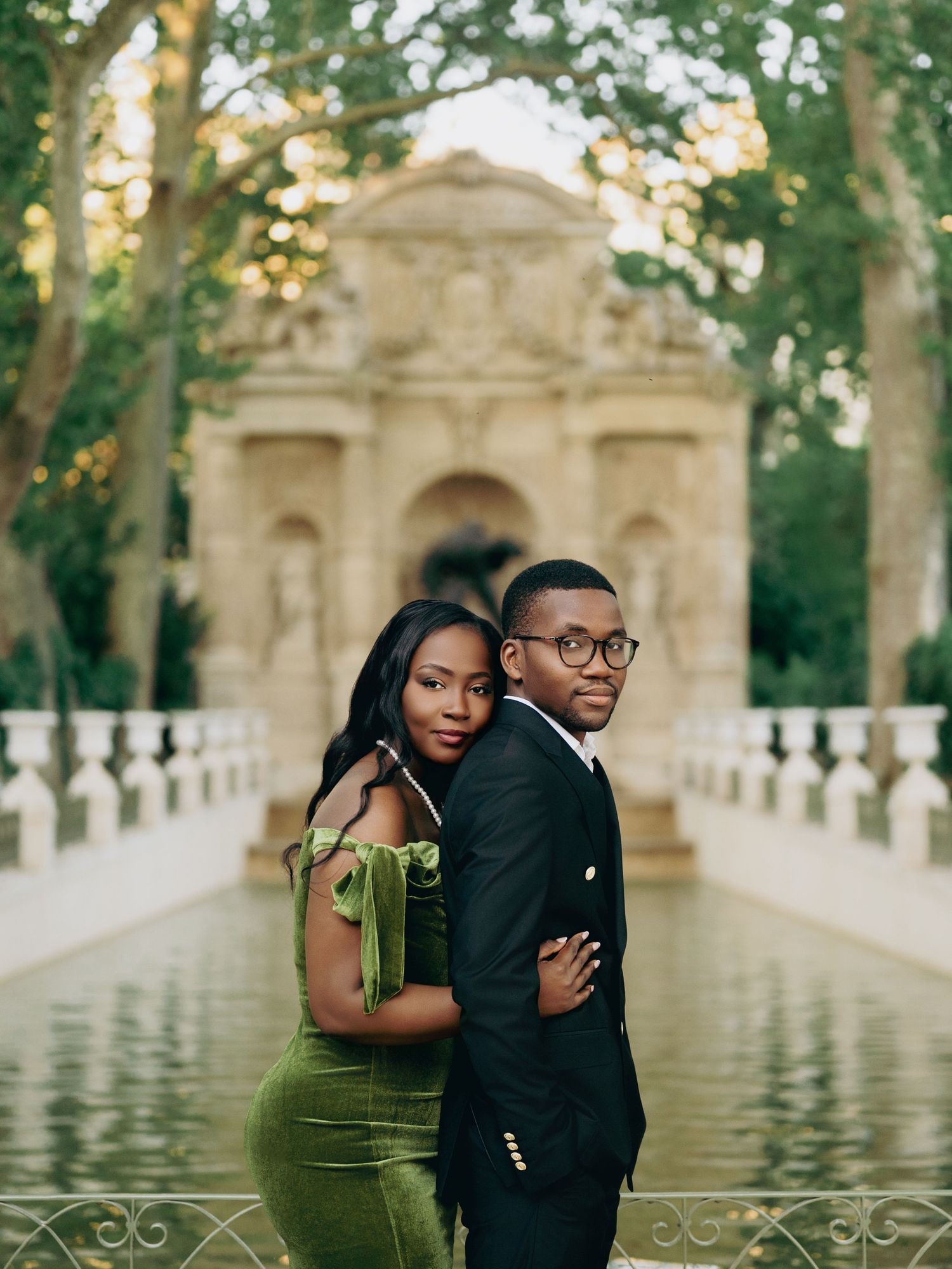 Sophisticated Black couple posing back-to-back before ornate stone fountain in lush garden setting.