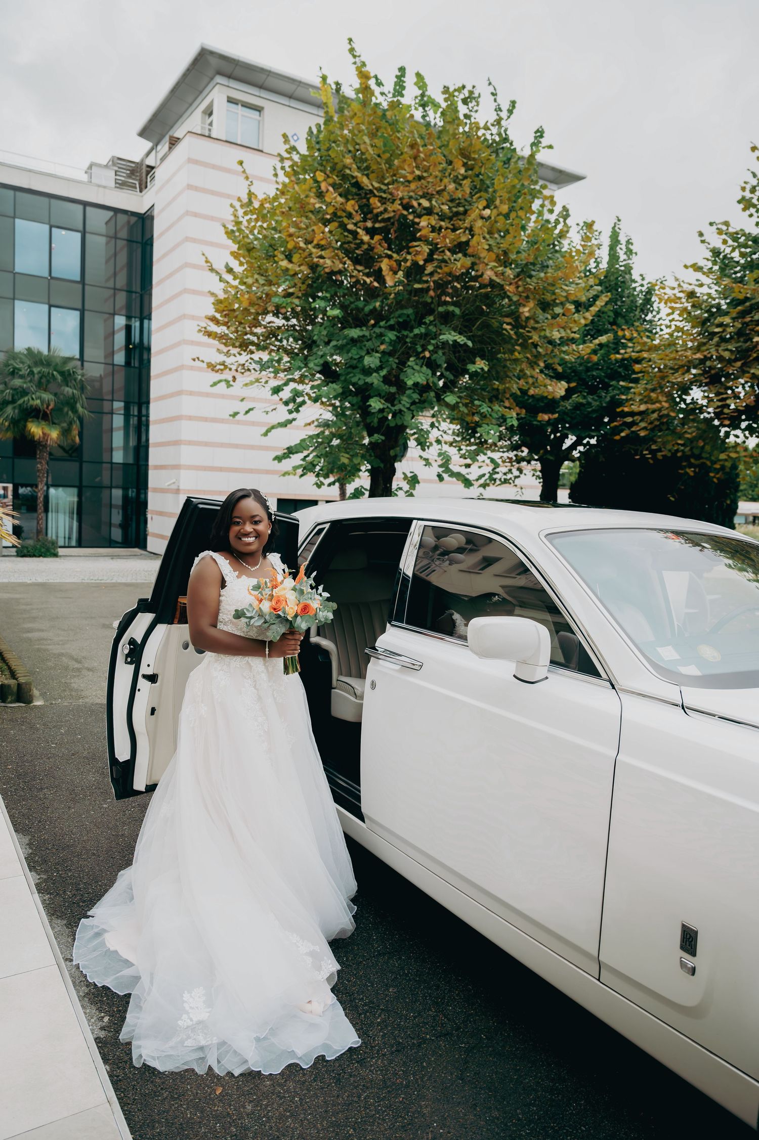 Smiling bride in white gown holding colorful bouquet stepping out of white Rolls-Royce wedding car.