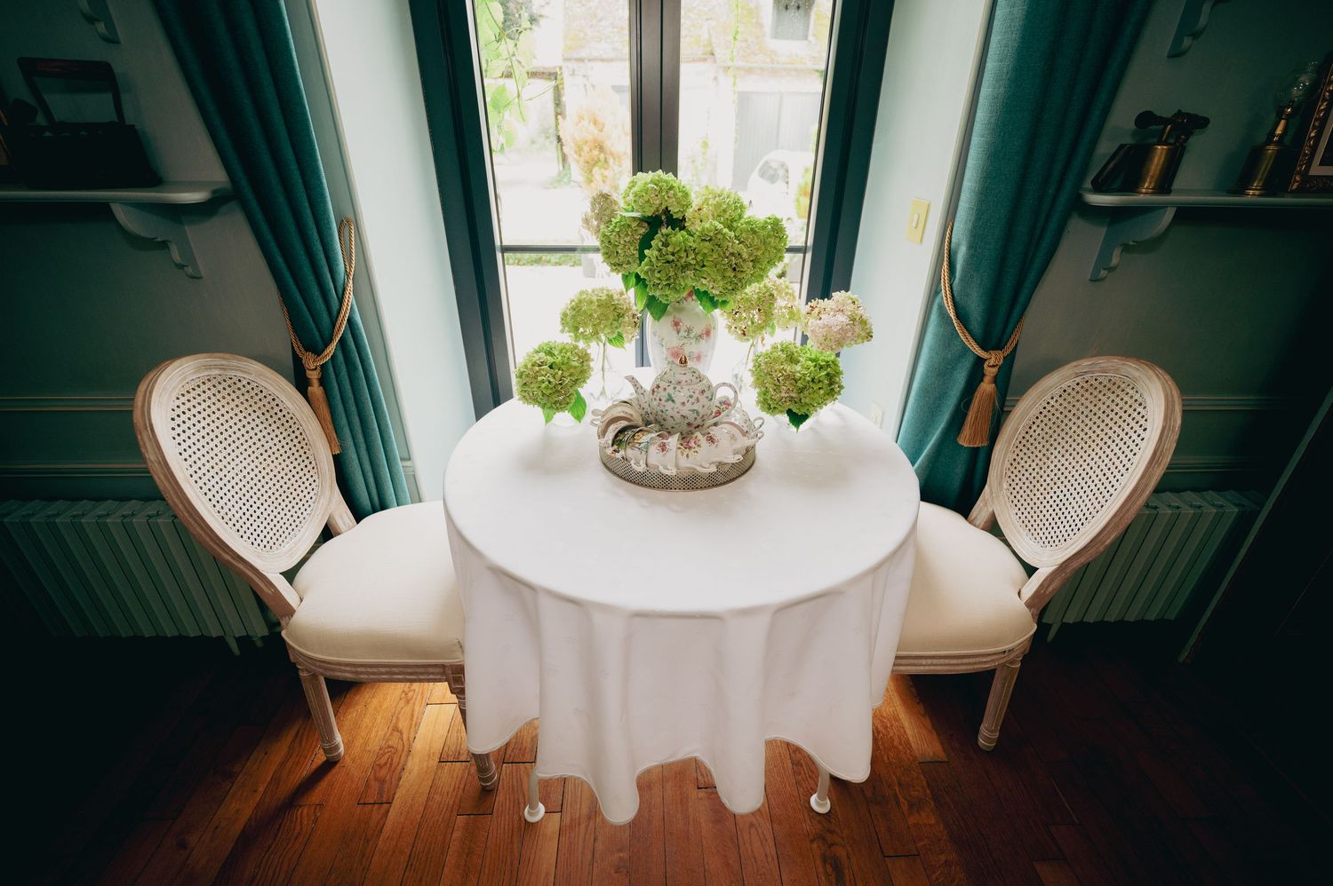 Elegant round table with white linen and green floral centerpiece flanked by two French chairs near teal curtained windows.