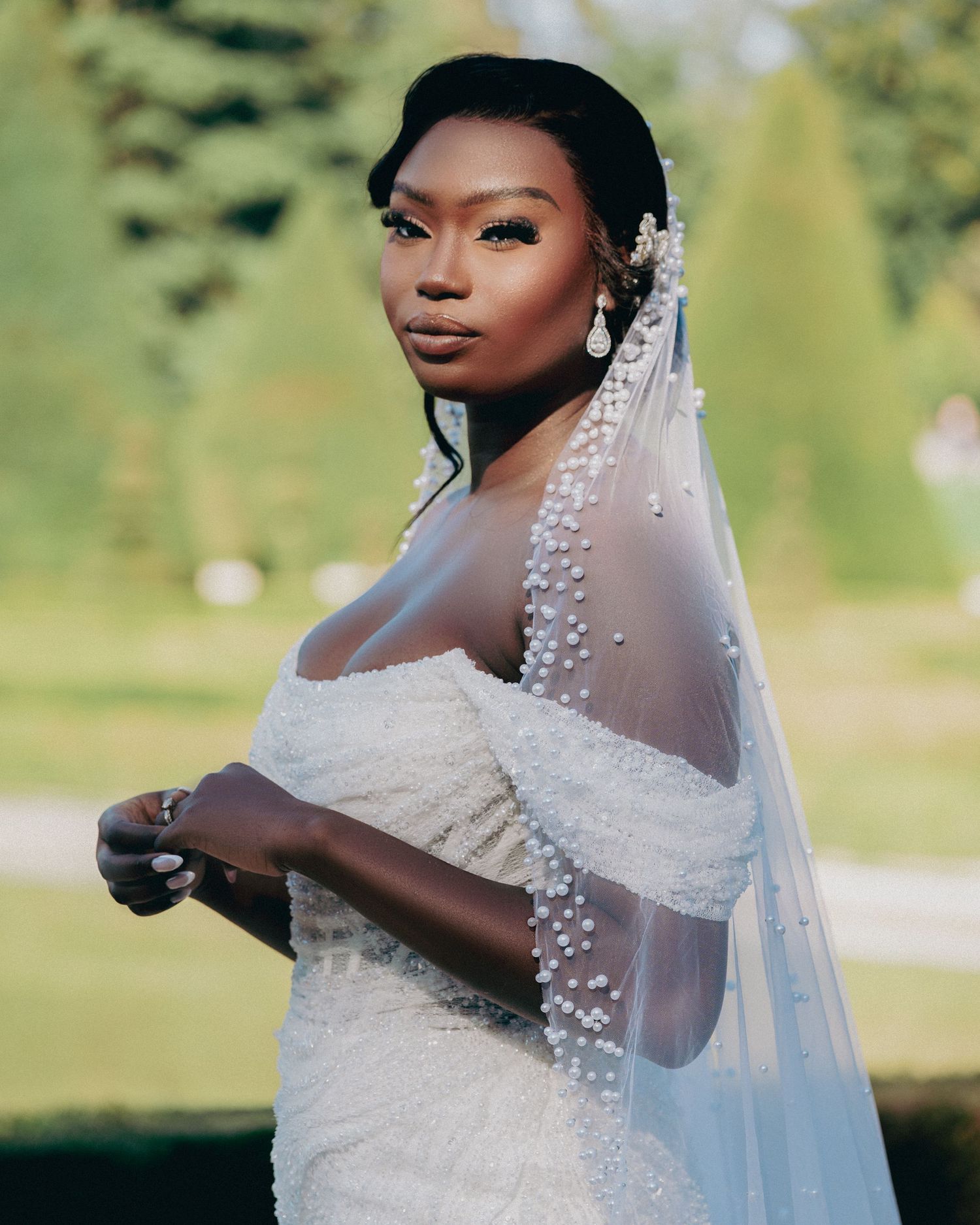Stunning Black bride in an off-shoulder beaded gown with a pearl-trimmed veil poses in a lush green garden setting.