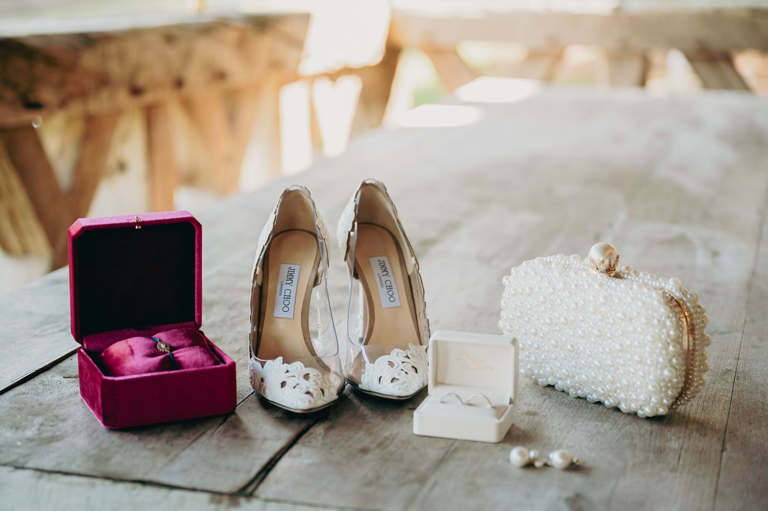 Wedding flat lay featuring white lace heels, pearl clutch, pink ring box, and accessories.