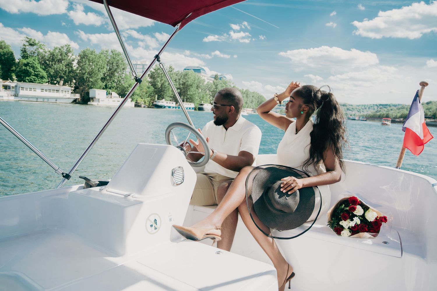 A stylish couple relaxes on a yacht deck, with a woman in white and a man steering near a French flag.