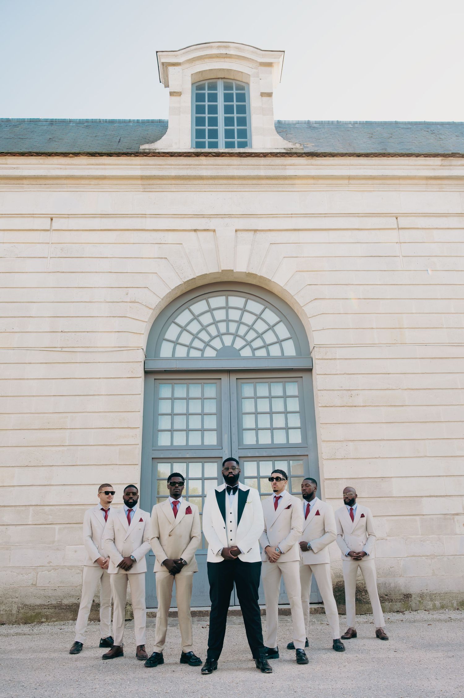 Groom in white tuxedo stands before groomsmen in cream suits outside a grand building.