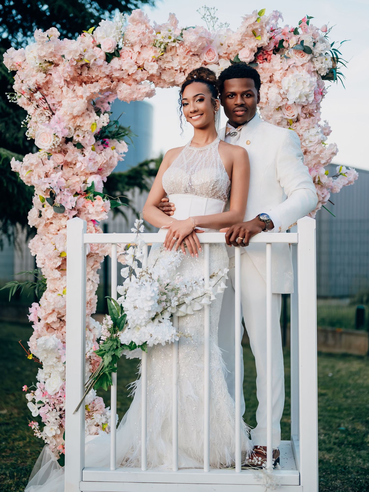 Bride and groom posing under a romantic pink floral arch, bride in lace gown holding white bouquet outdoors.