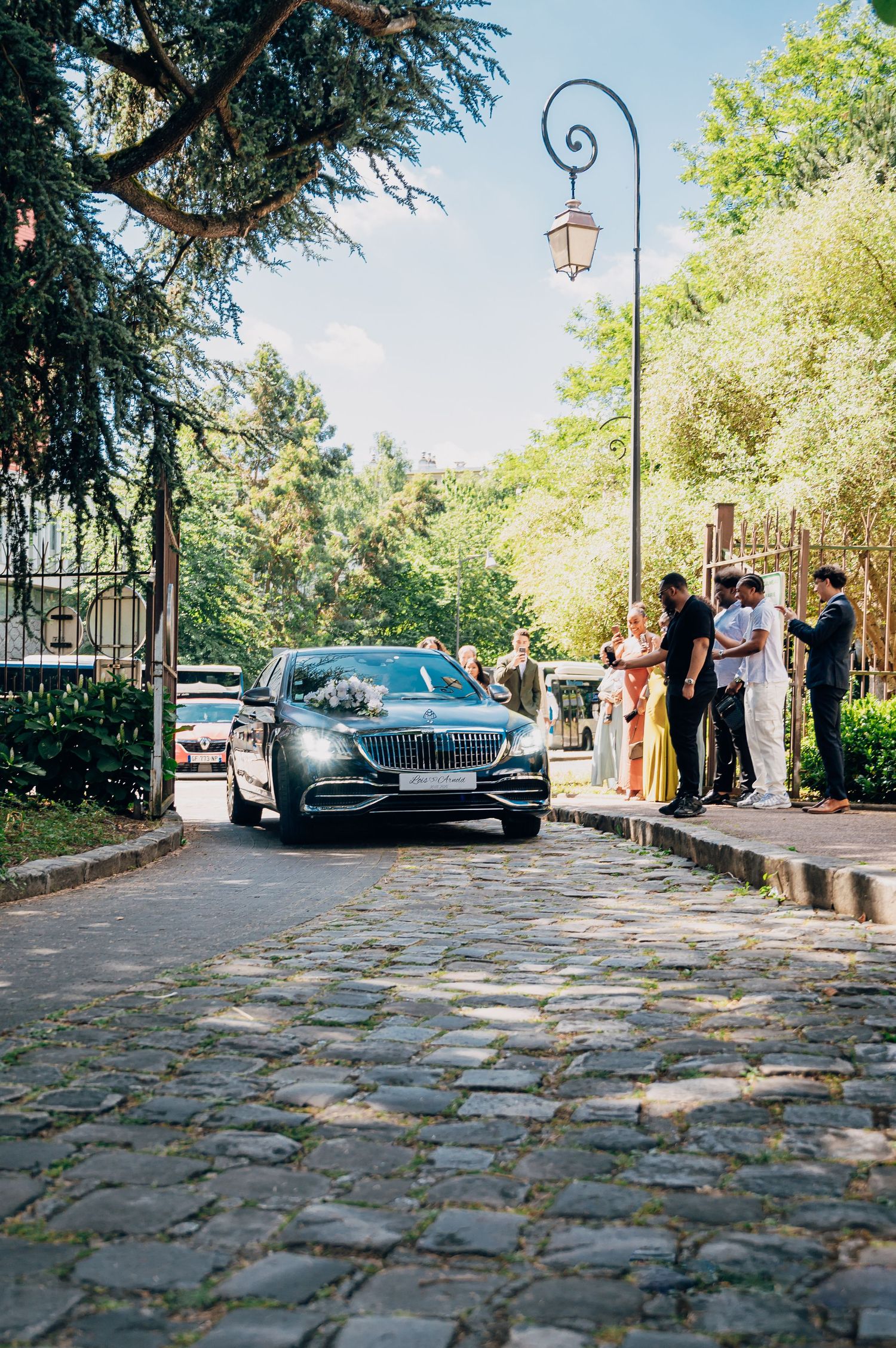 Luxury black Mercedes-Maybach wedding car arriving on cobblestone driveway with guests.
