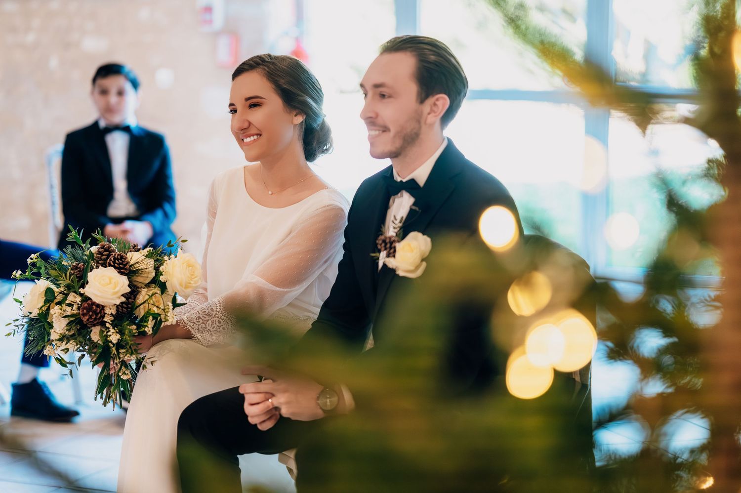 Smiling bride and groom holding hands during winter wedding ceremony framed by Christmas tree lights.