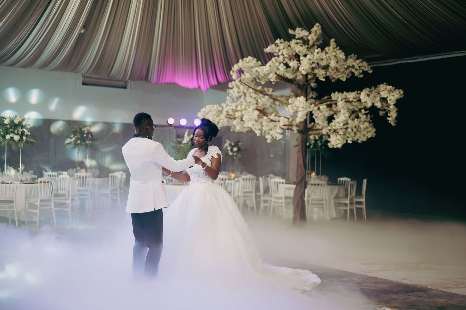 Couple shares romantic first dance amid misty atmosphere near stunning white cherry blossom tree.