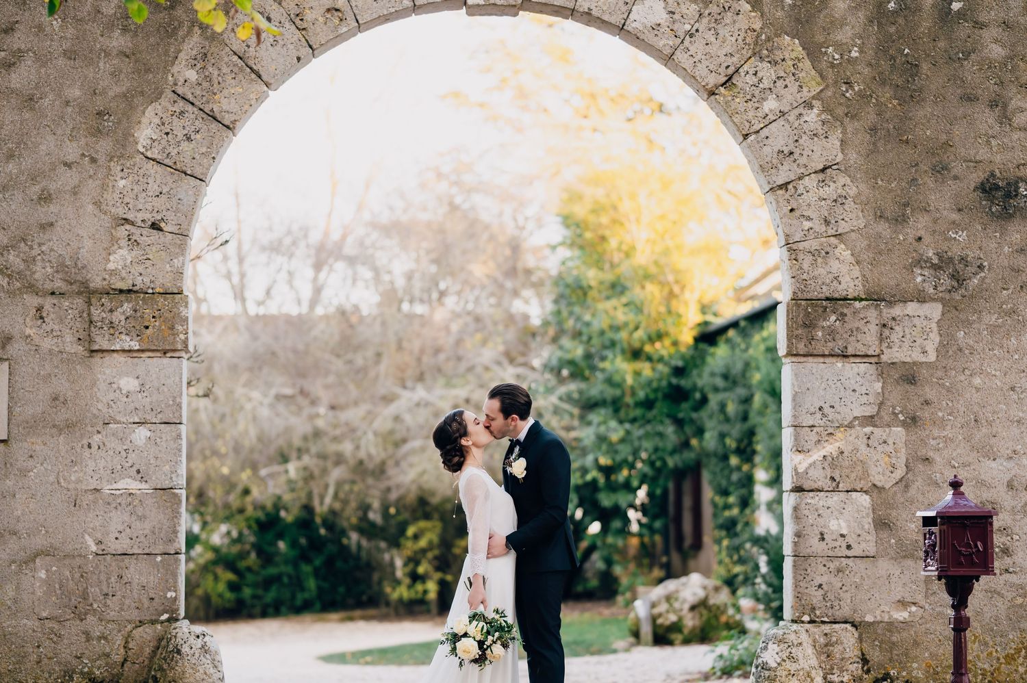 Bride and groom share a romantic kiss under a stone archway surrounded by autumn foliage at their wedding.