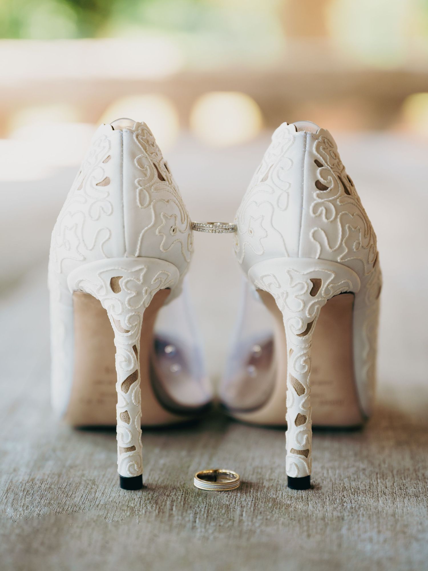 Elegant white laser-cut bridal heels with gold wedding rings placed between them on a soft surface.