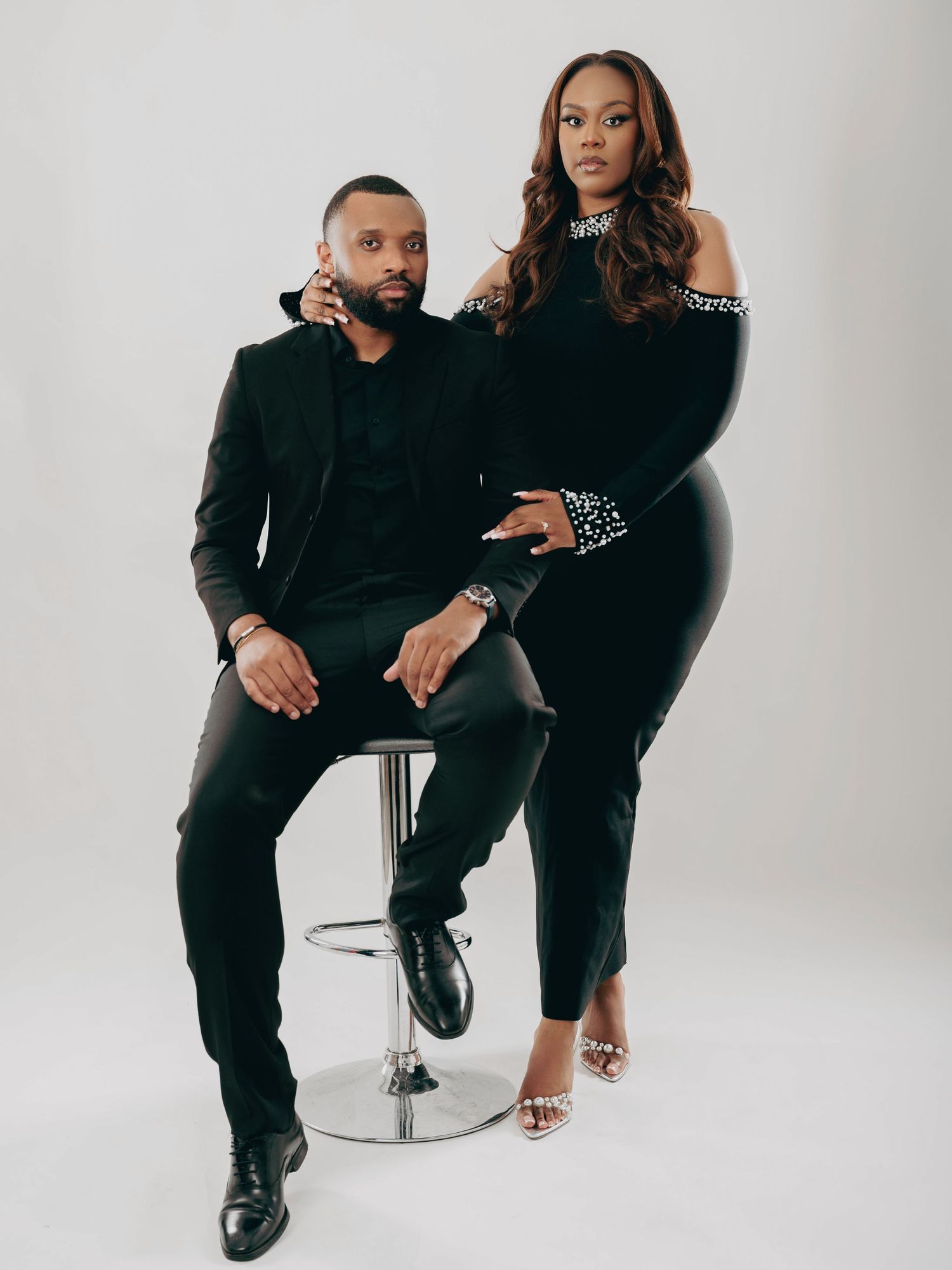 Stylish couple dressed in all black posing together on a chrome barstool against a white background.